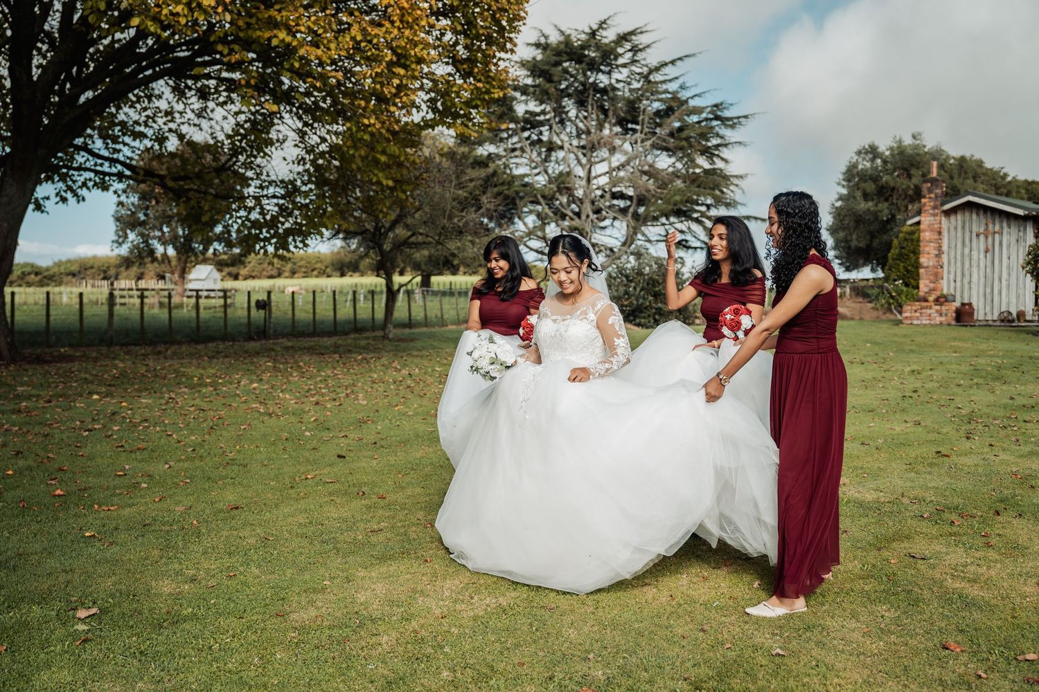 Bride in white gown with bridesmaids in burgundy dresses helping carry her train on green lawn.
