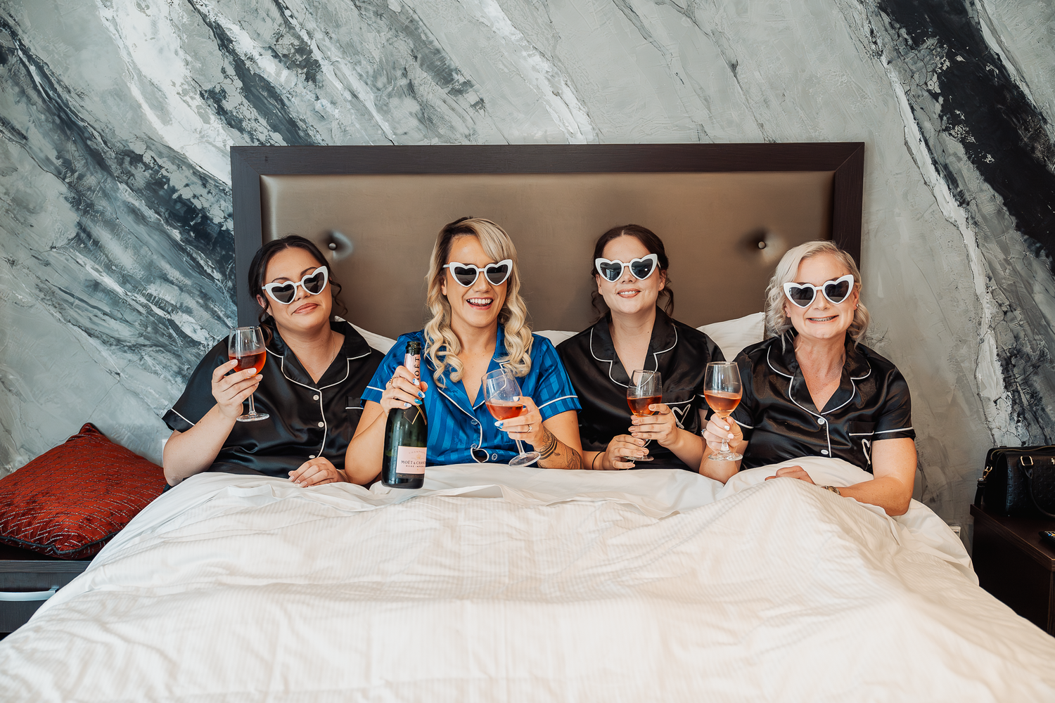 Four women in sunglasses lounging on bed during wedding preparations.