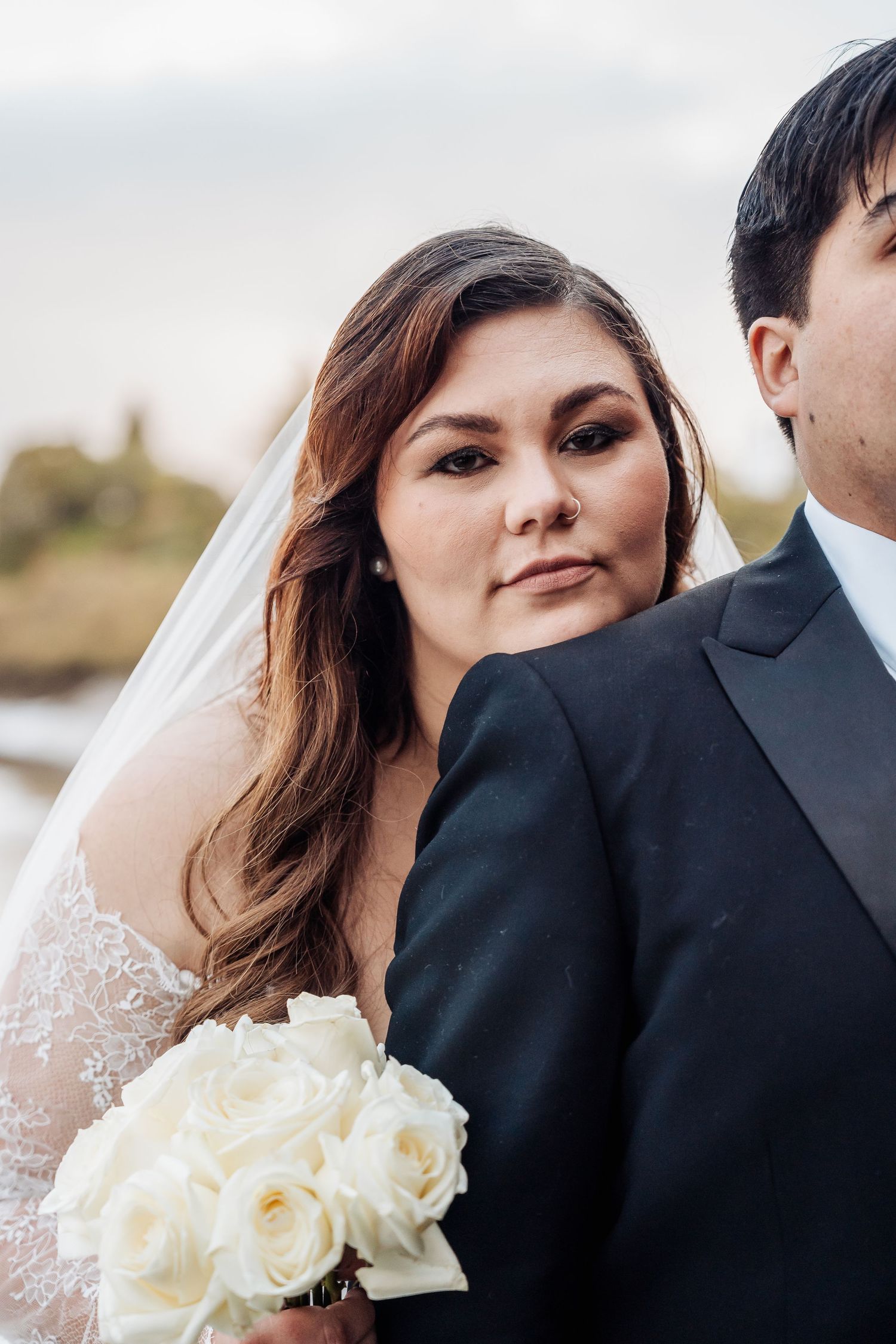 A bride in lace holds a bouquet of white roses while standing next to her groom in a black suit on their wedding day.