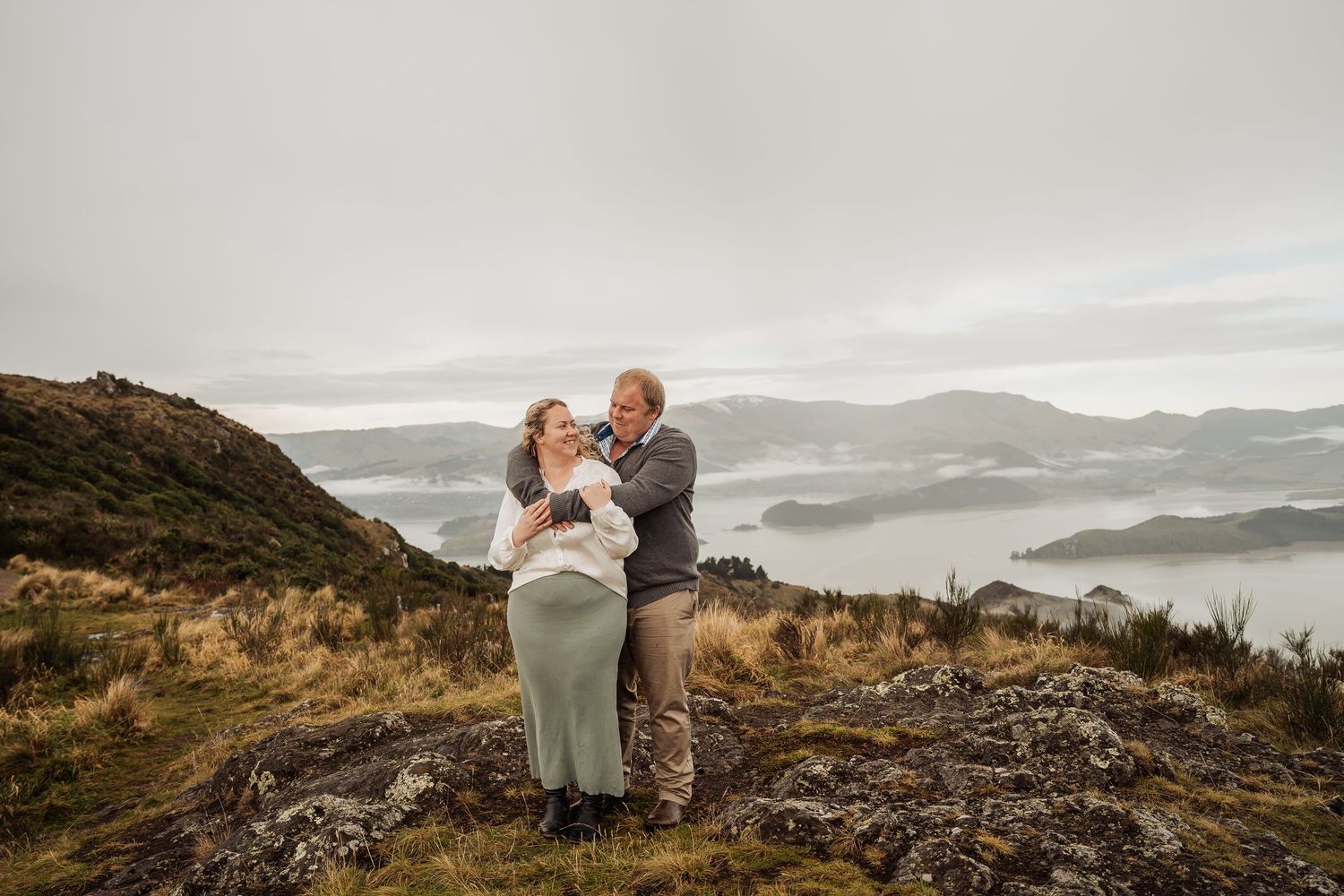 A couple shares an intimate moment on a mountaintop overlooking misty valleys and rolling hills at sunset.