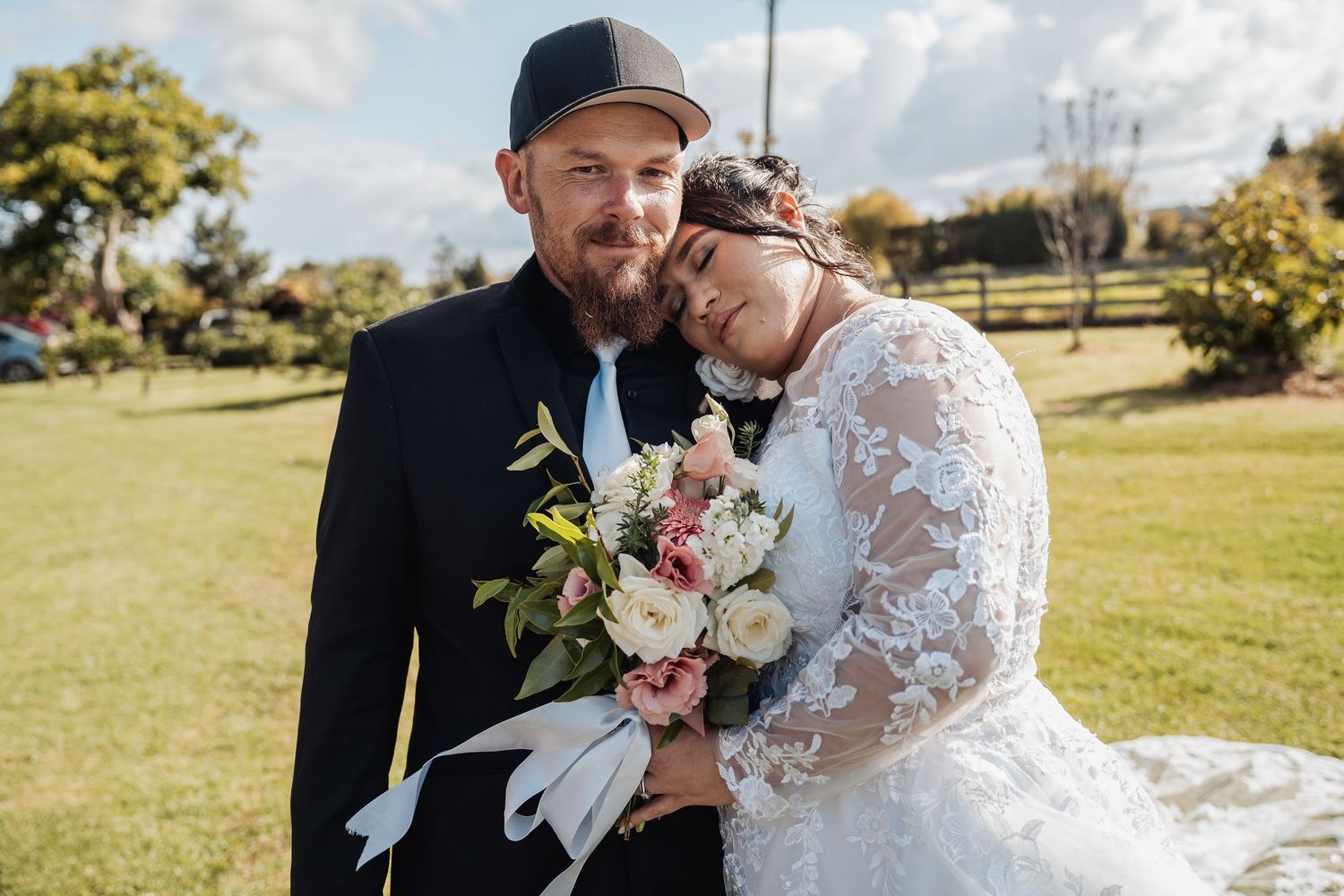 A happy couple shares an intimate moment in a sunny garden setting while holding a beautiful floral bouquet.
