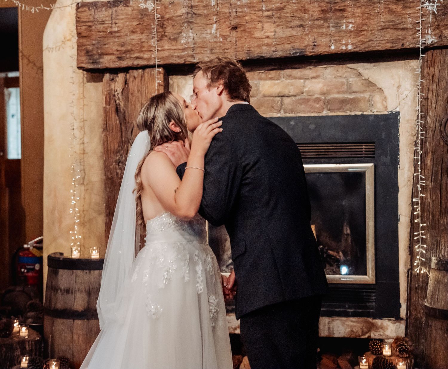 A couple shares their first kiss during a romantic wedding ceremony in a rustic brick venue with a fireplace.
