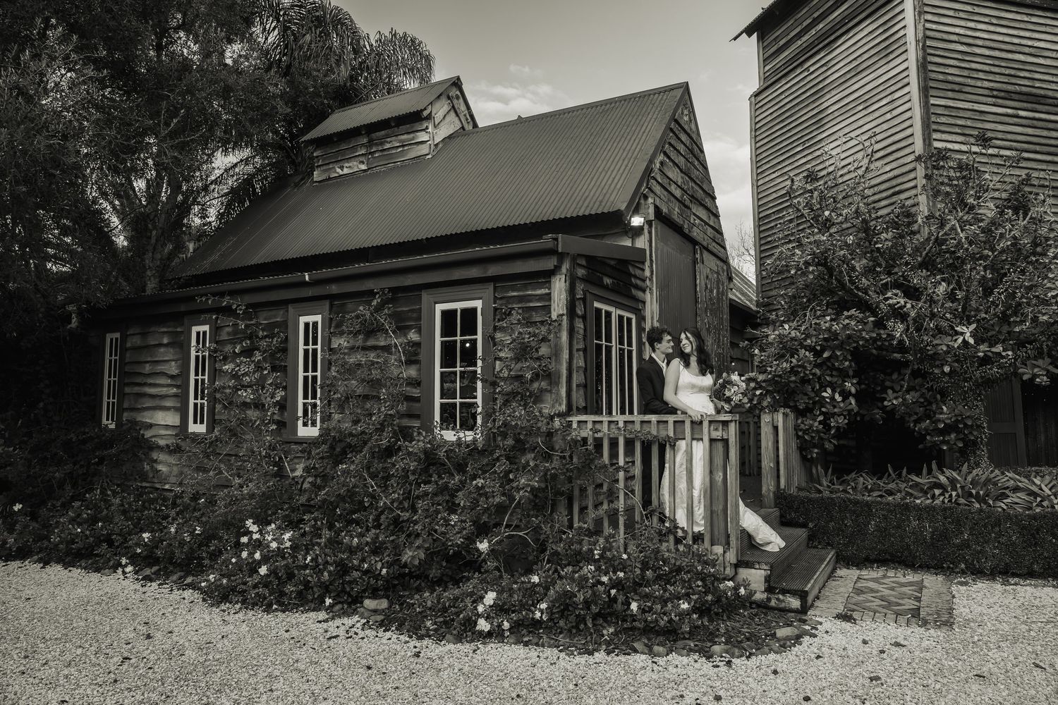 Black and white photo of a couple posing outside a rustic stone cottage surrounded by lush gardens.
