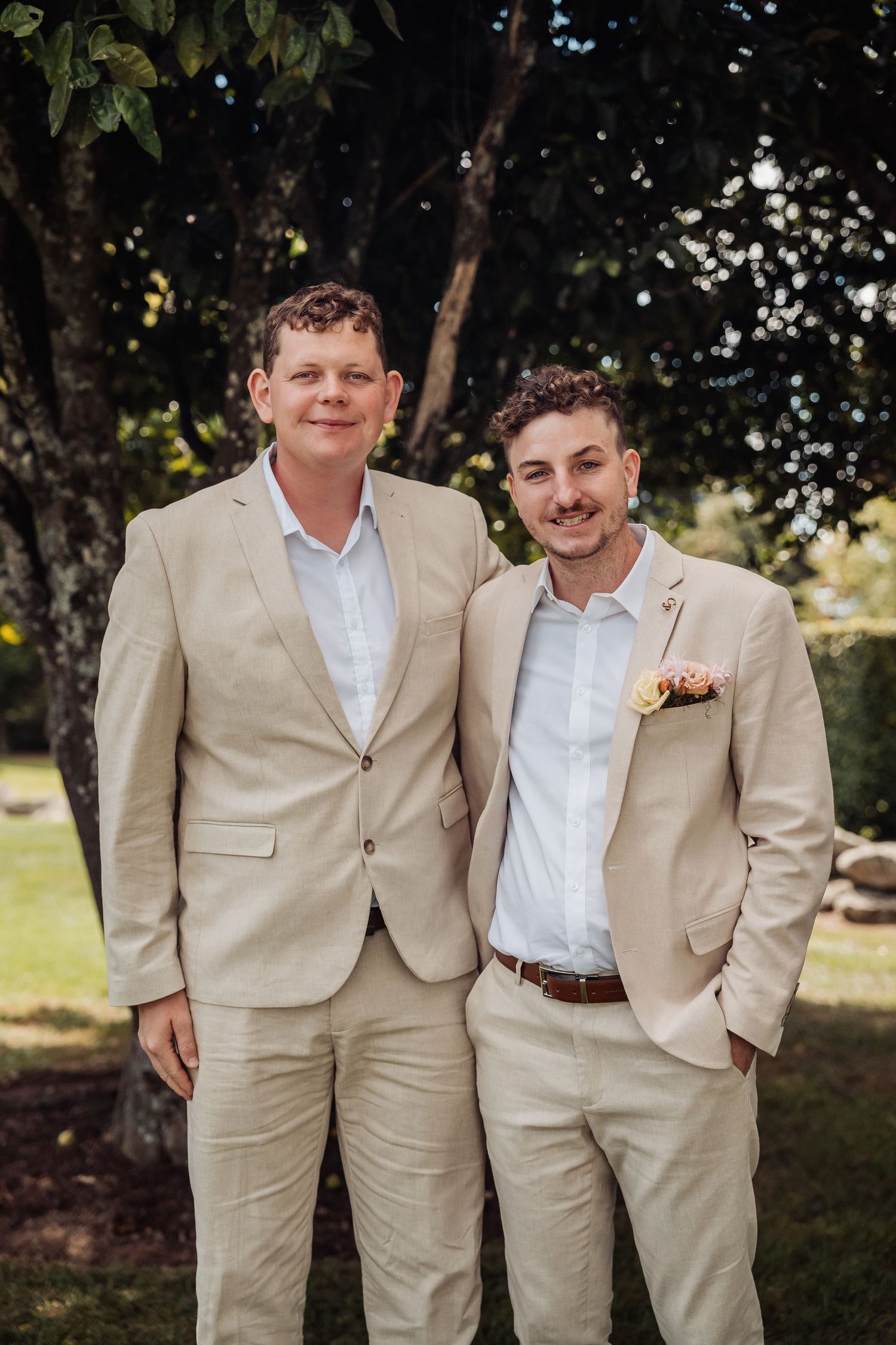Two groomsmen smiling in matching beige suits standing outdoors under green trees.
