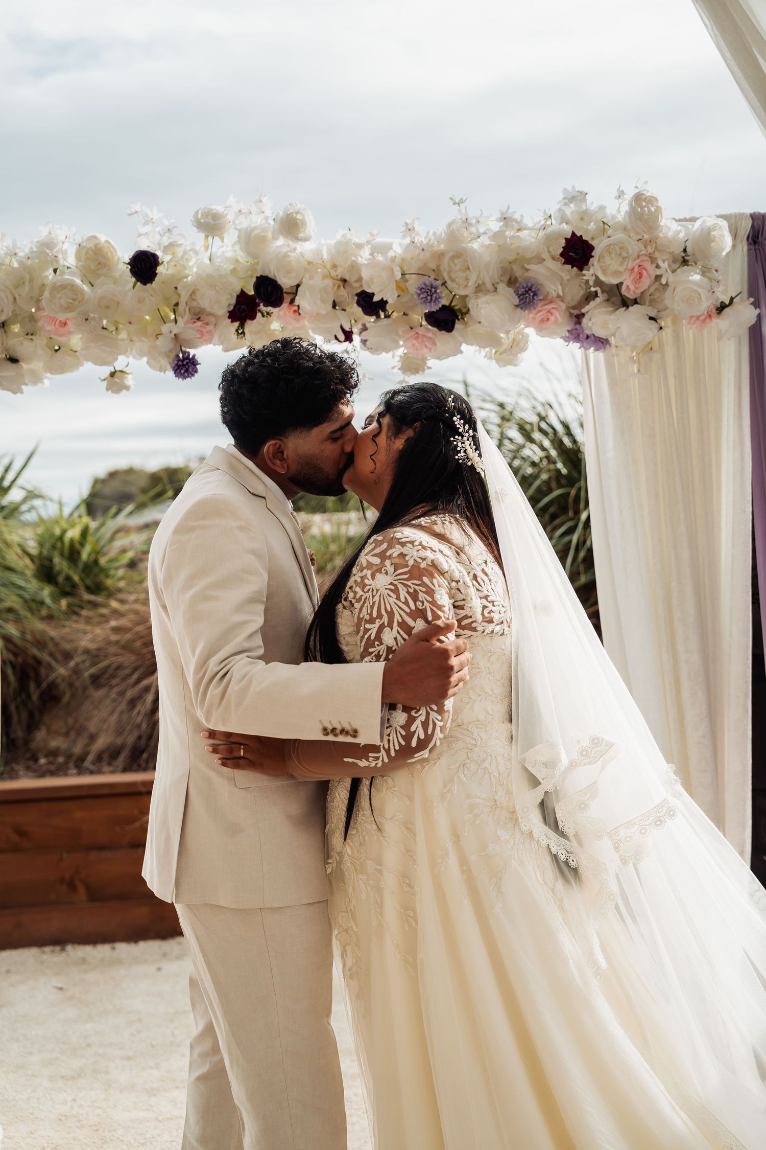 Bride and groom sharing first kiss under floral arch at outdoor wedding ceremony.