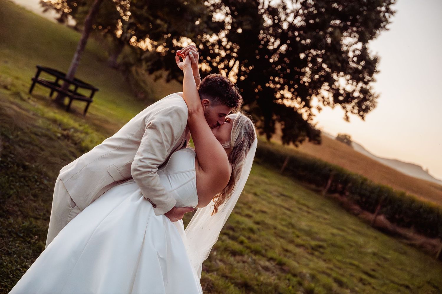 Groom dips bride romantically outdoors at sunset during wedding photo session.