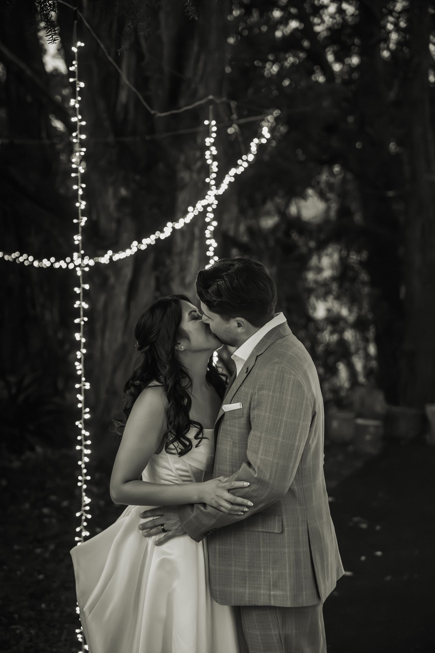 Black and white photo of bride and groom kissing beneath twinkling fairy lights in forest.