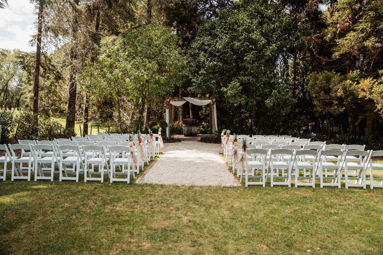 Outdoor wedding ceremony setup with white chairs, gravel aisle, and rustic floral arch amid lush trees.