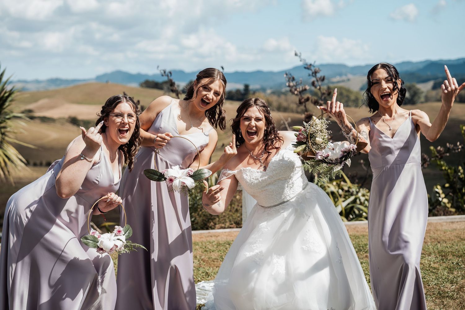 Bride in white gown celebrating joyfully with three bridesmaids in lavender dresses against a scenic hillside.