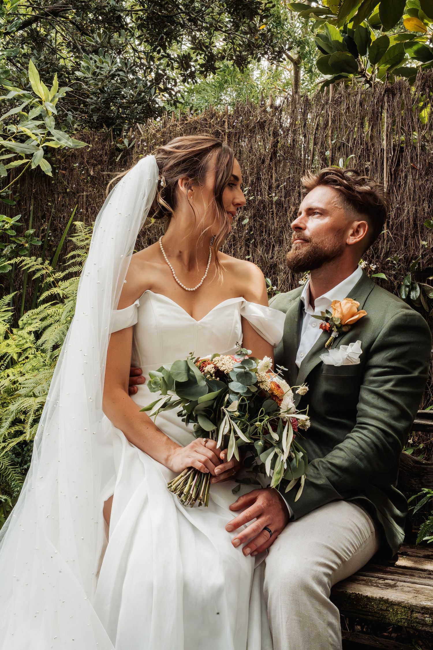 Bride and groom gazing at each other on a bench in a tropical garden, bride holding bouquet.