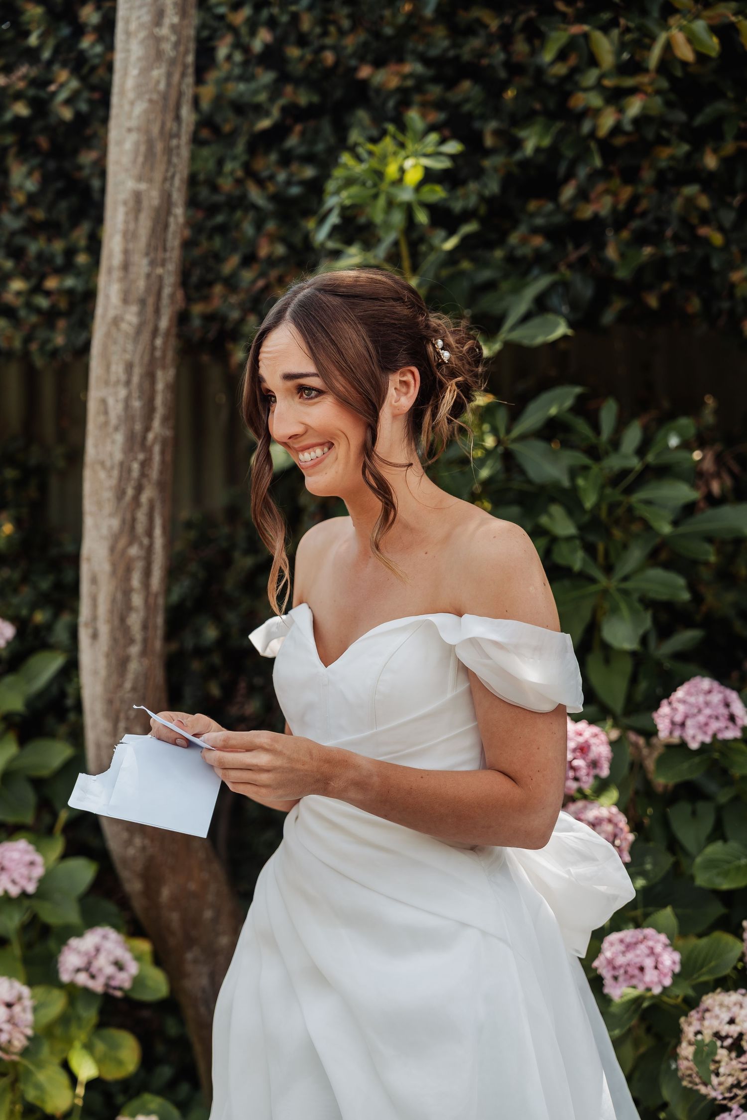Radiant bride in elegant off-shoulder wedding dress holding vow cards among blooming hydrangeas.