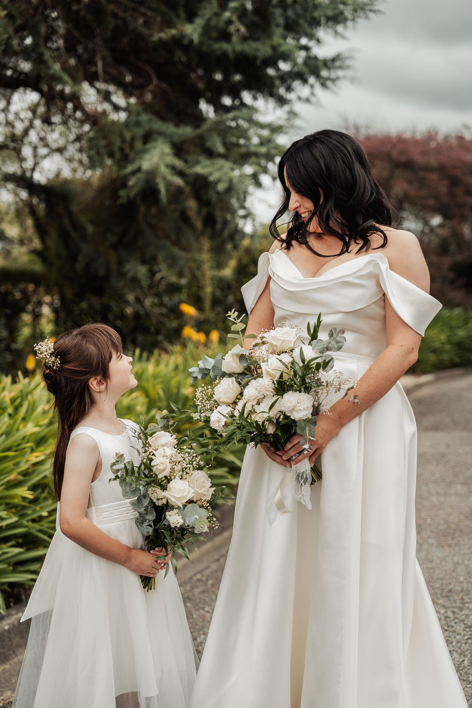 Bride in off-shoulder white gown smiles at flower girl holding bouquets in a garden.