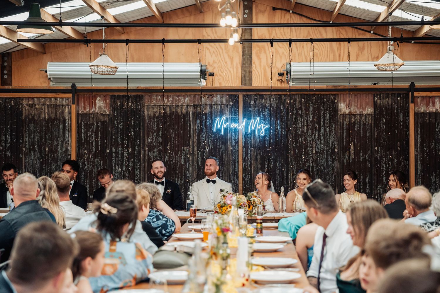 Bride and groom at head table during wedding reception in a decorated barn with a neon Mr and Mrs sign.