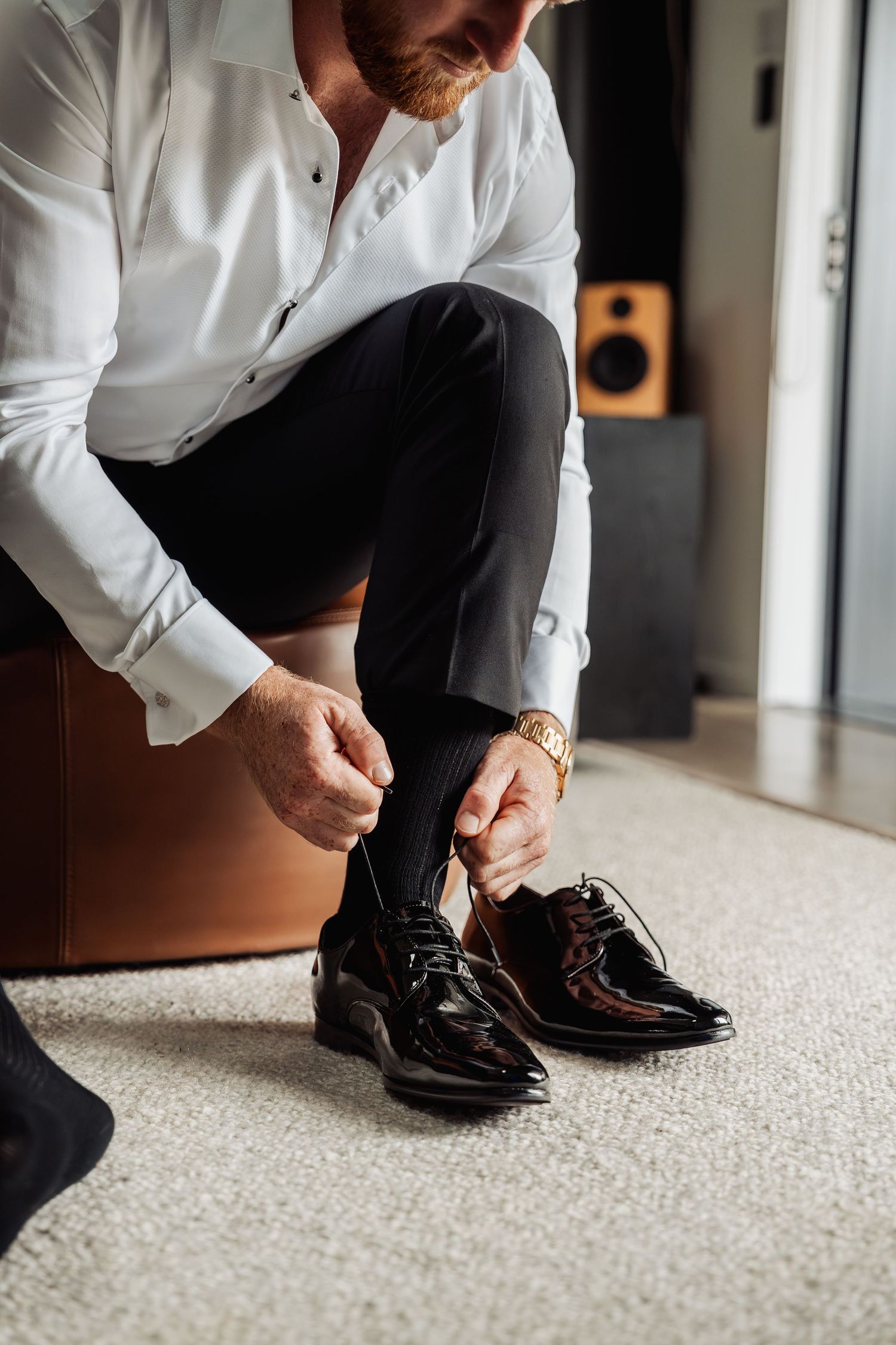 Groom in white shirt tying black patent leather shoes while wearing a gold watch on wedding day.