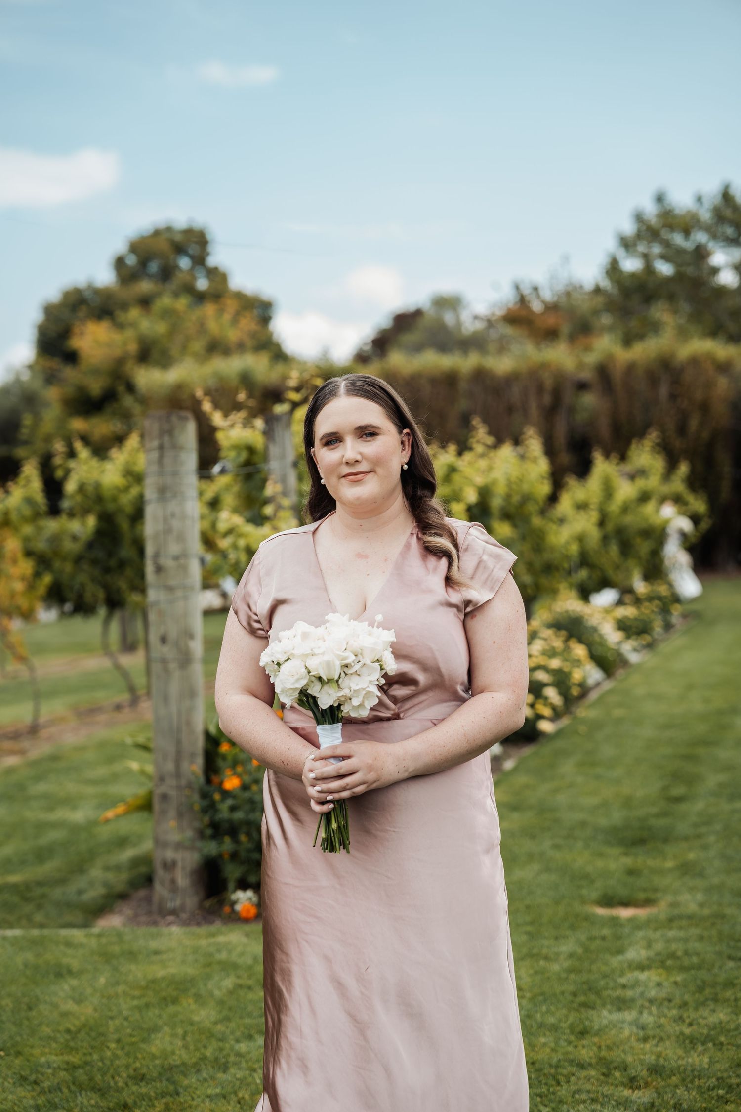 Bridesmaid in dusty pink dress holds white floral bouquet while standing in a lush garden vineyard setting.