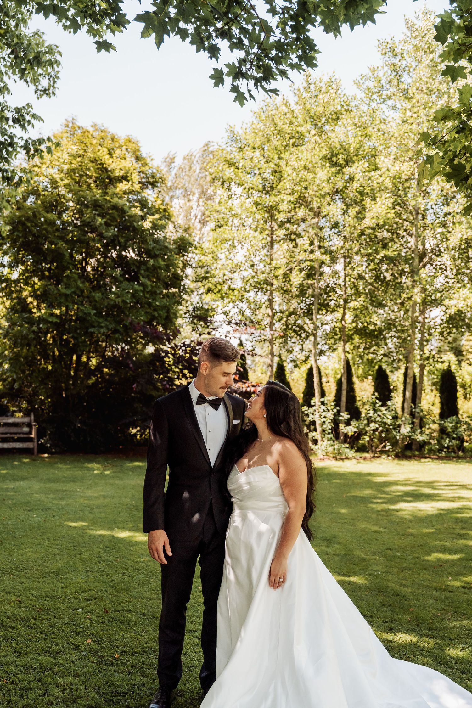 Bride and groom share a romantic moment in a lush green garden, bride in white satin gown, groom in black tuxedo.