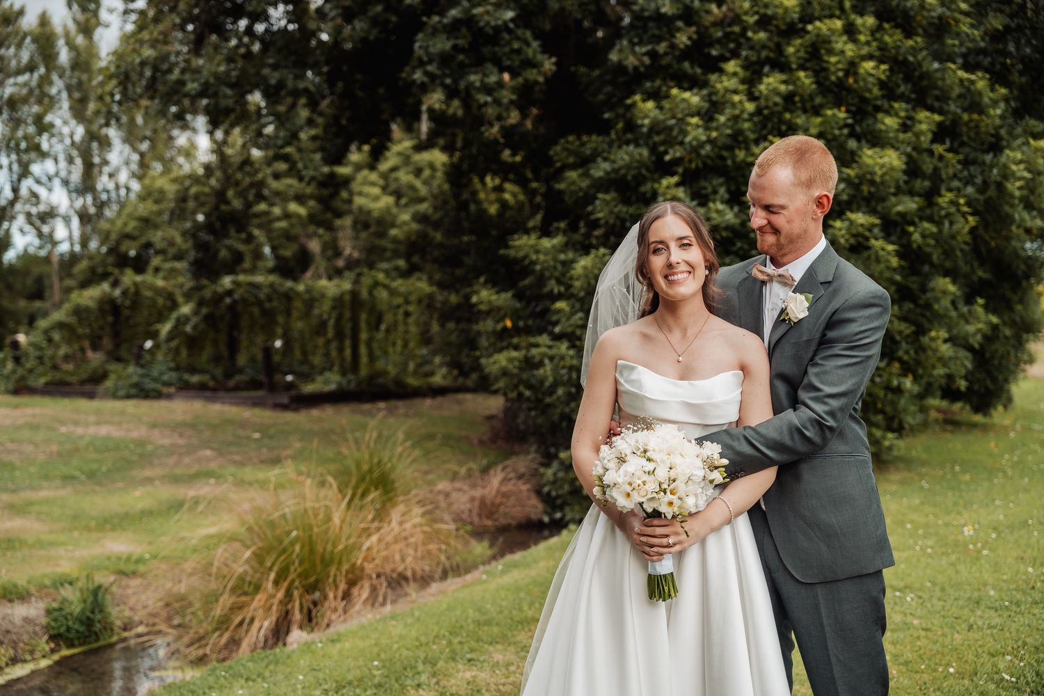 Bride and groom embracing in a lush garden setting, bride holding a white bouquet in elegant wedding attire.
