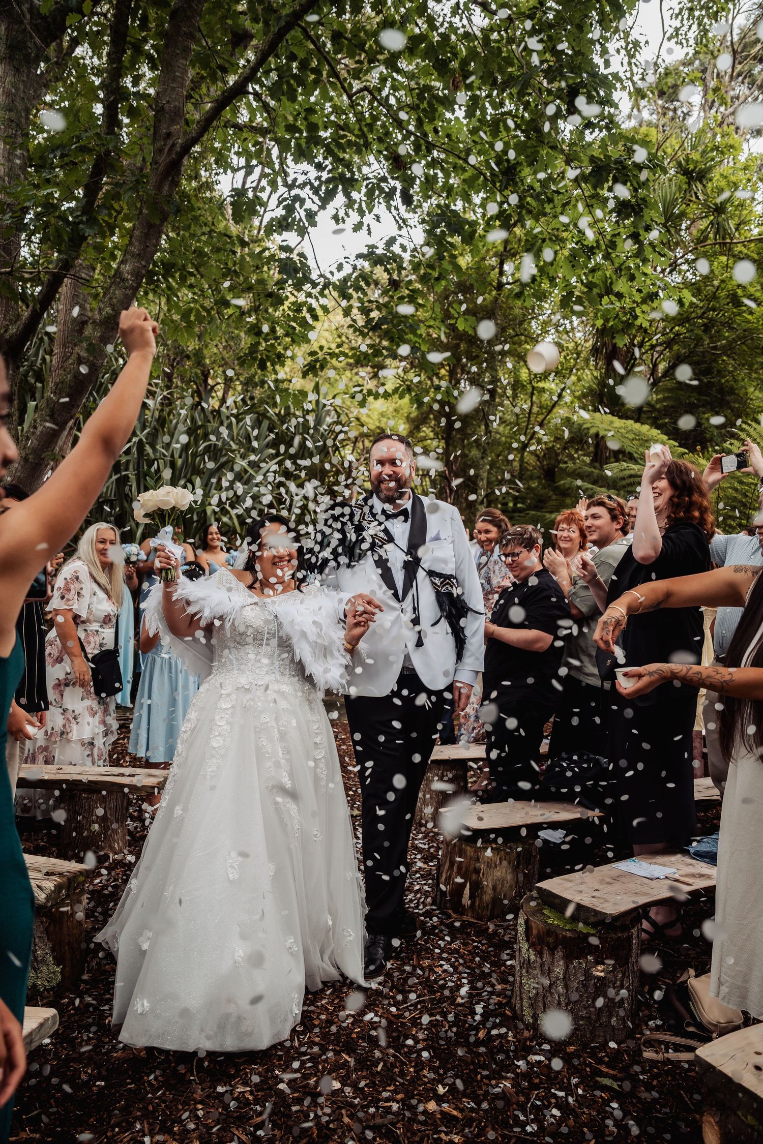 Newlyweds walk through confetti shower at joyful outdoor forest wedding reception.
