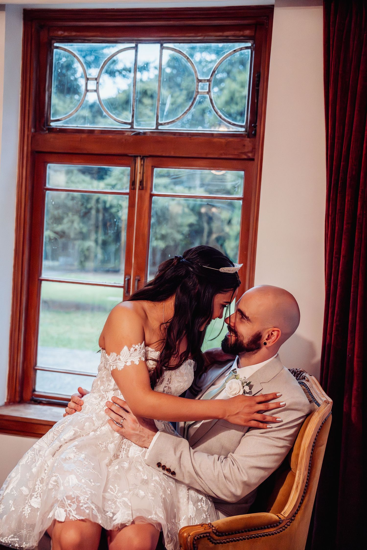 Bride in lace gown sitting on groom's lap sharing a tender moment by a stained glass window.