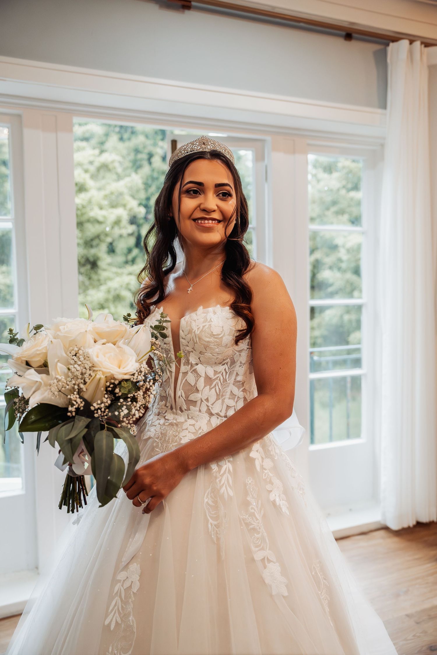 Smiling bride in strapless lace gown holding white bouquet wearing a tiara.