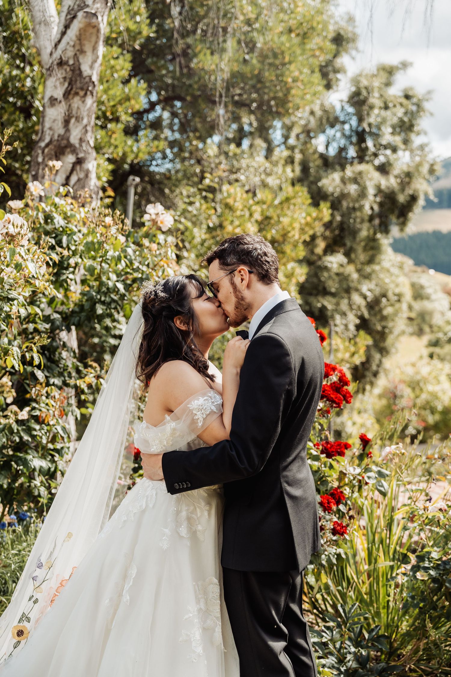 Bride and groom share romantic kiss surrounded by colorful roses in lush garden setting.