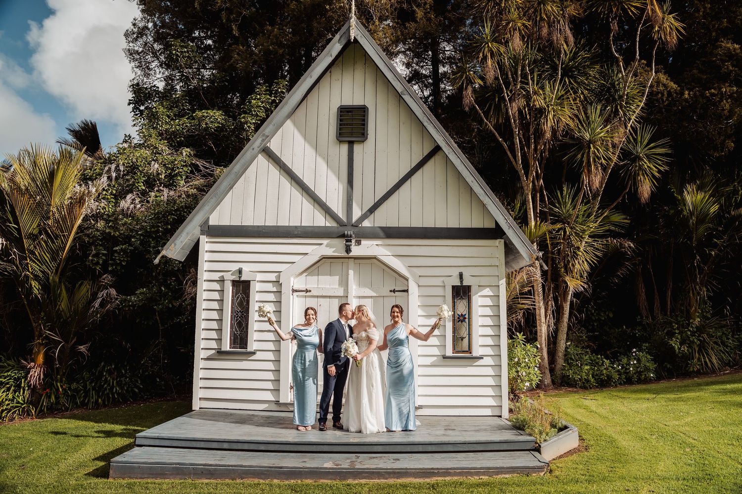 Small white chapel with cross sits nestled among palm trees and greenery.