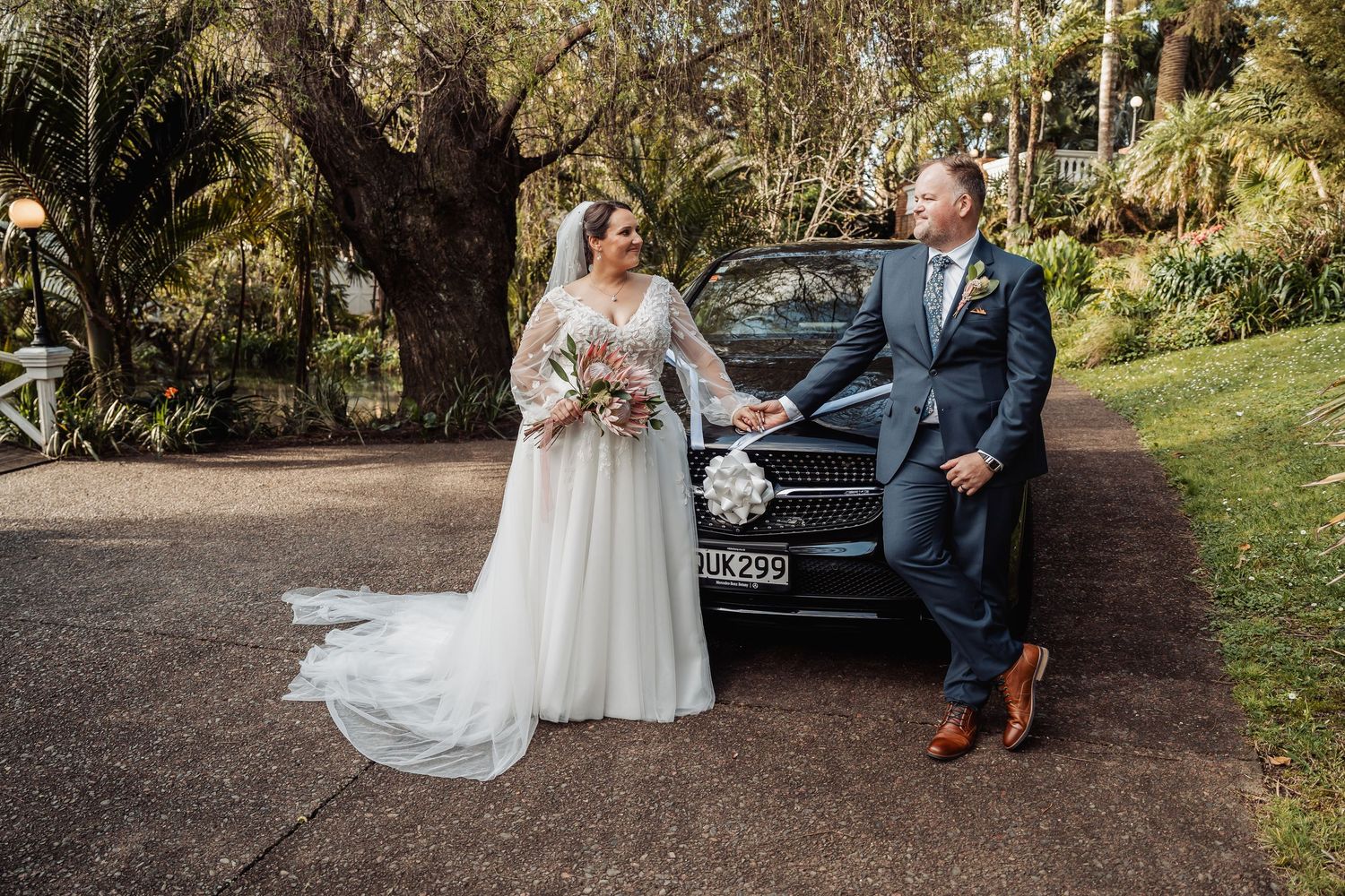 A couple poses together outdoors near a vintage car during a wedding photoshoot in a lush garden setting.