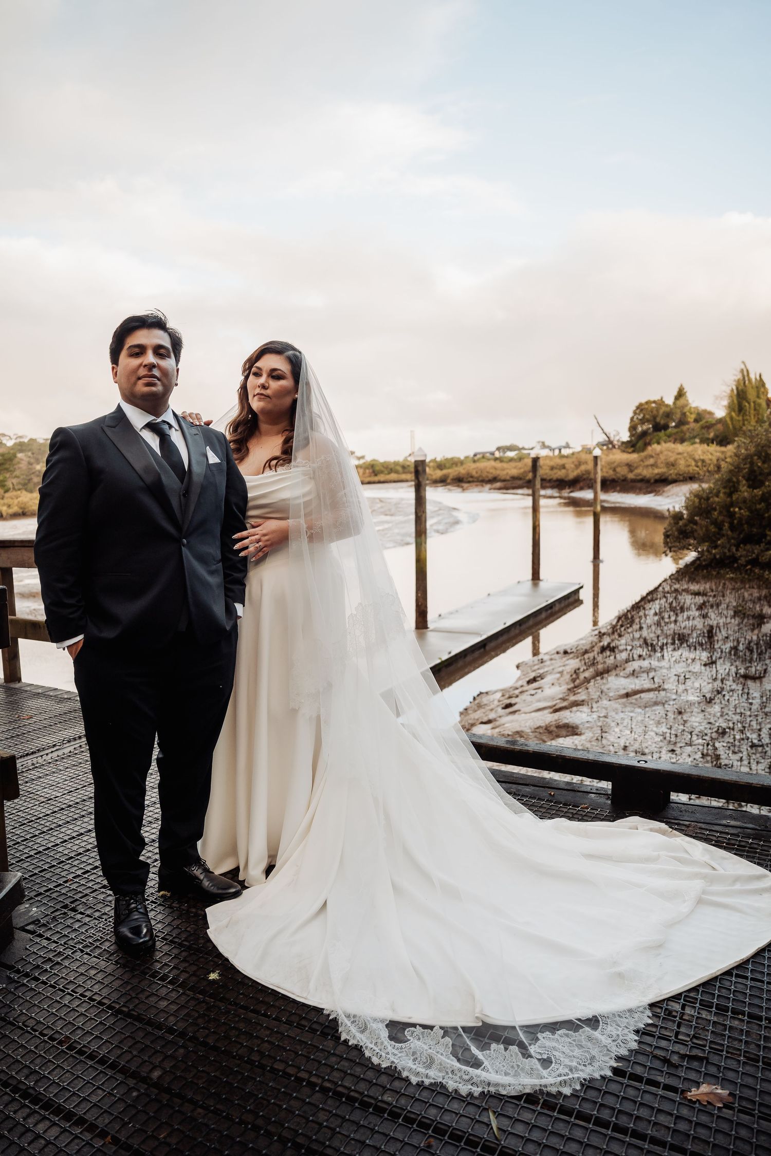 Elegant newlyweds pose together on a wooden dock by a serene waterway during an outdoor wedding photoshoot.