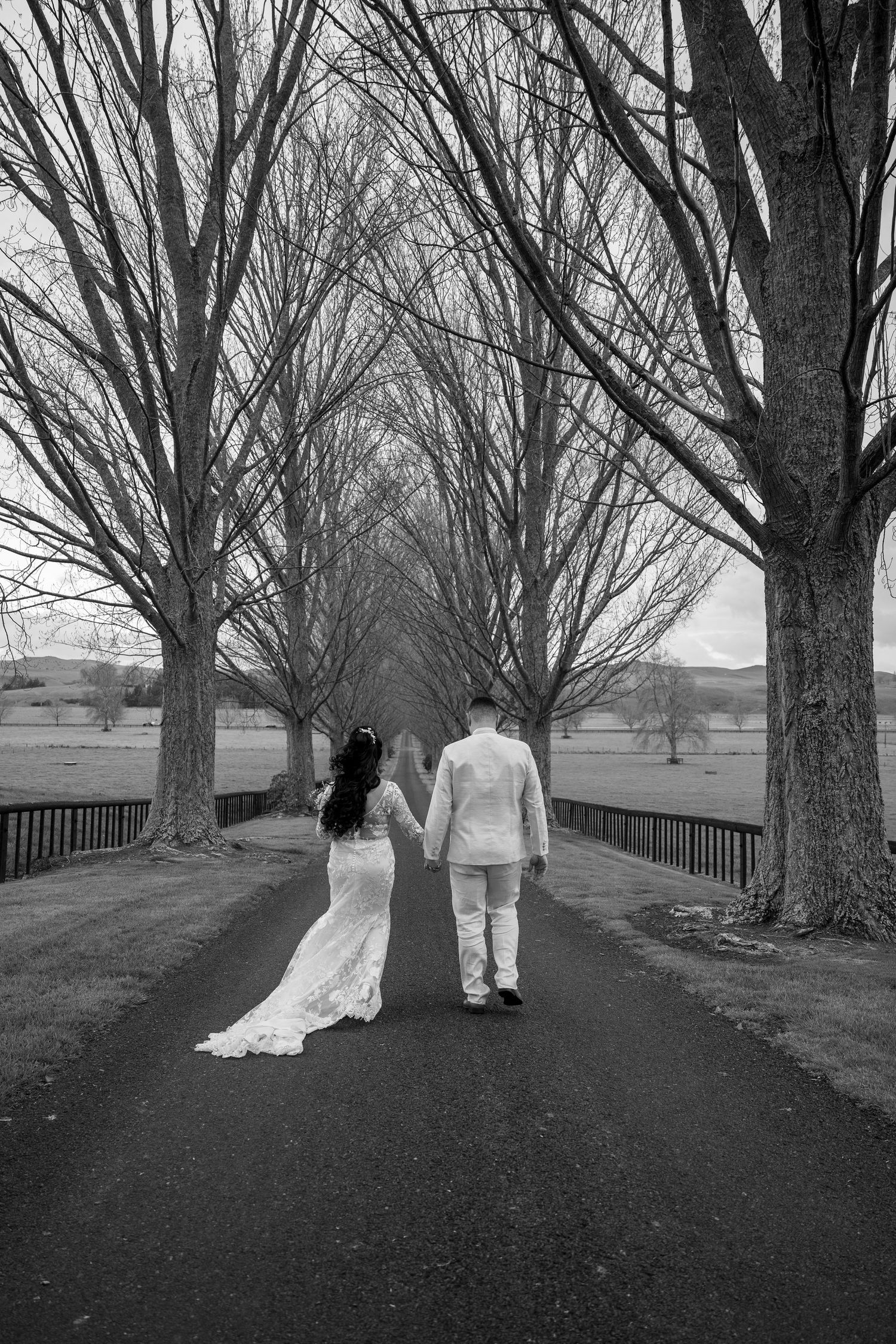 Black and white photo of newlyweds walking along a dramatic tree-lined path.