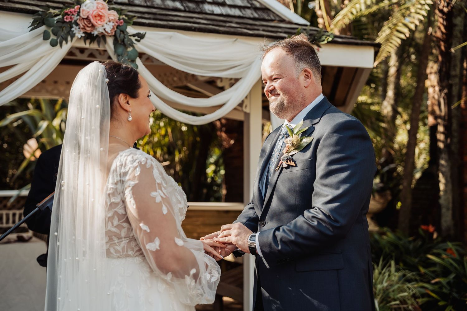 A couple shares a tender moment during their wedding ceremony under a white draped canopy in a garden setting.