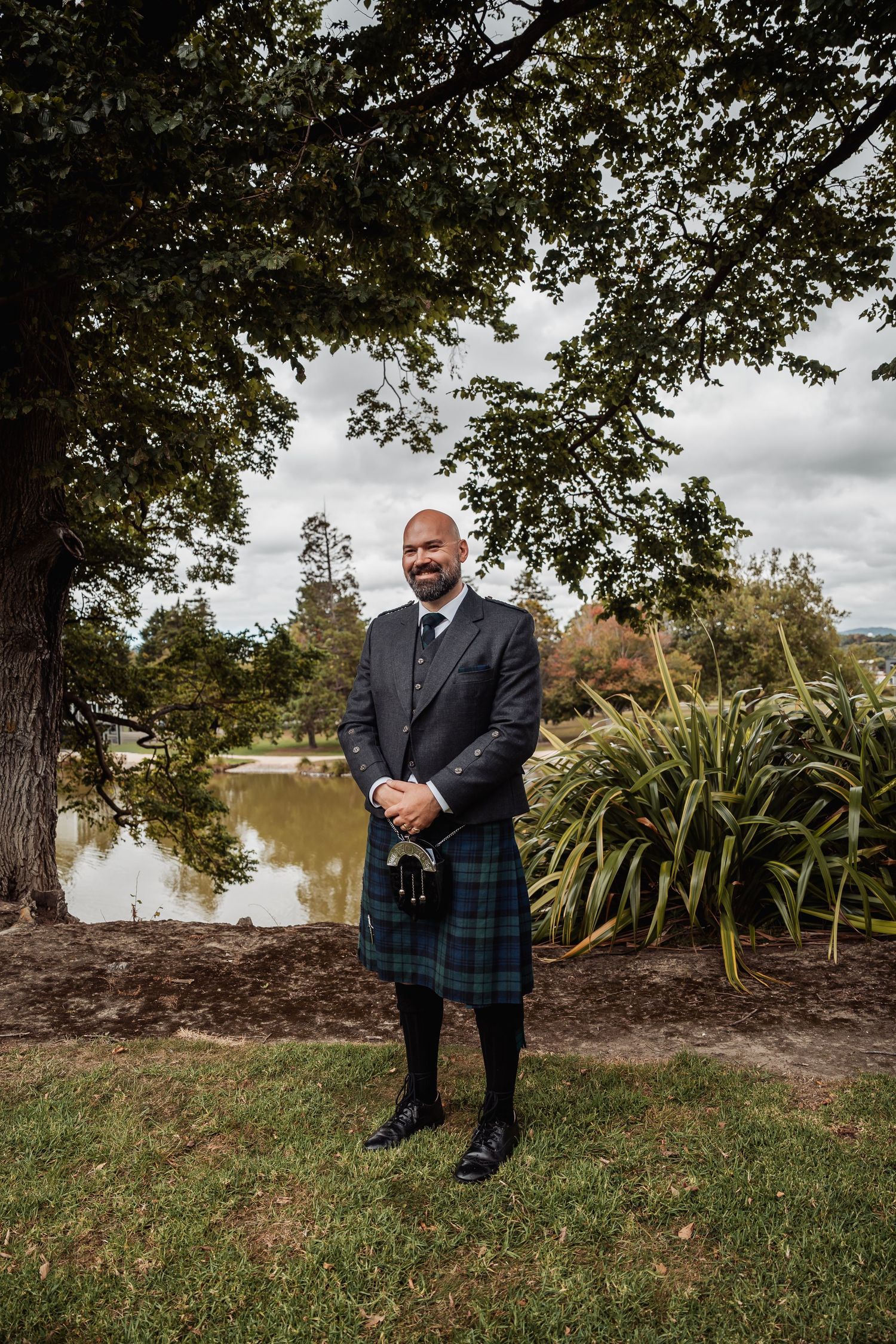 Groom in Scottish tartan kilt and jacket stands under large tree beside a scenic pond.