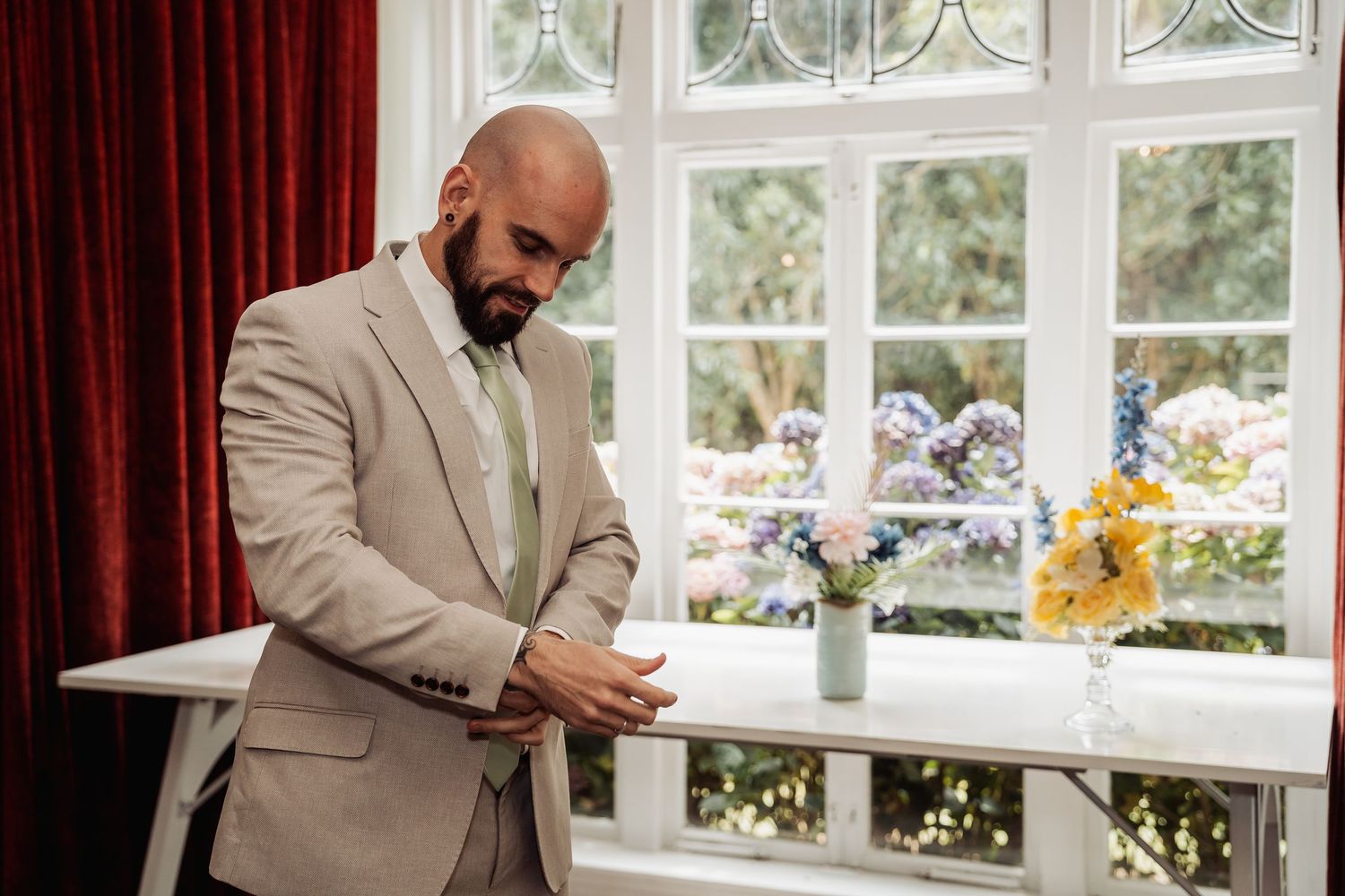 Groom in a beige suit adjusting his cufflinks near a window with flowers.