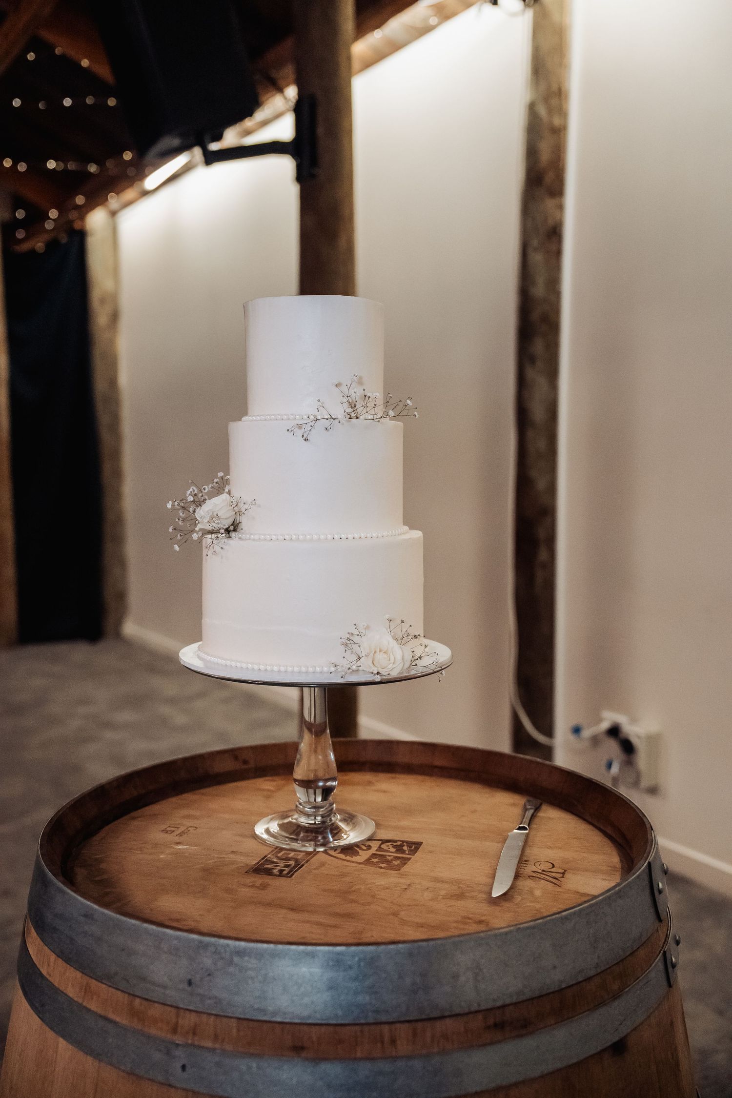 Elegant three-tier white wedding cake adorned with baby's breath, displayed on a rustic wooden barrel with a crystal cake stand.