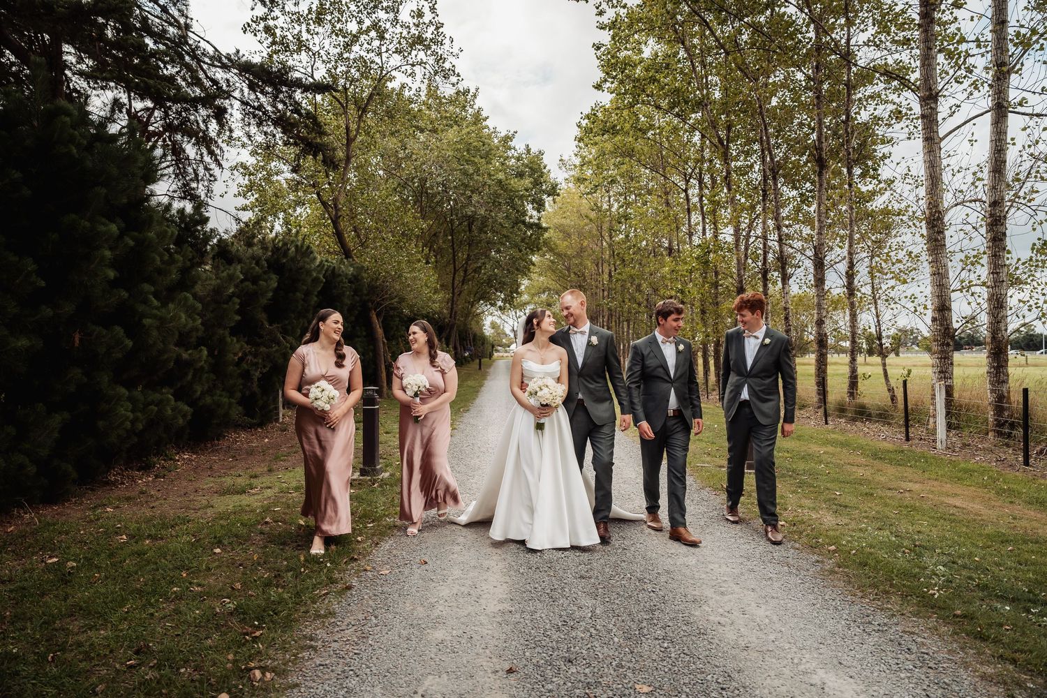 Wedding party walks together down a tree-lined gravel path with bridesmaids in blush and groomsmen in gray.