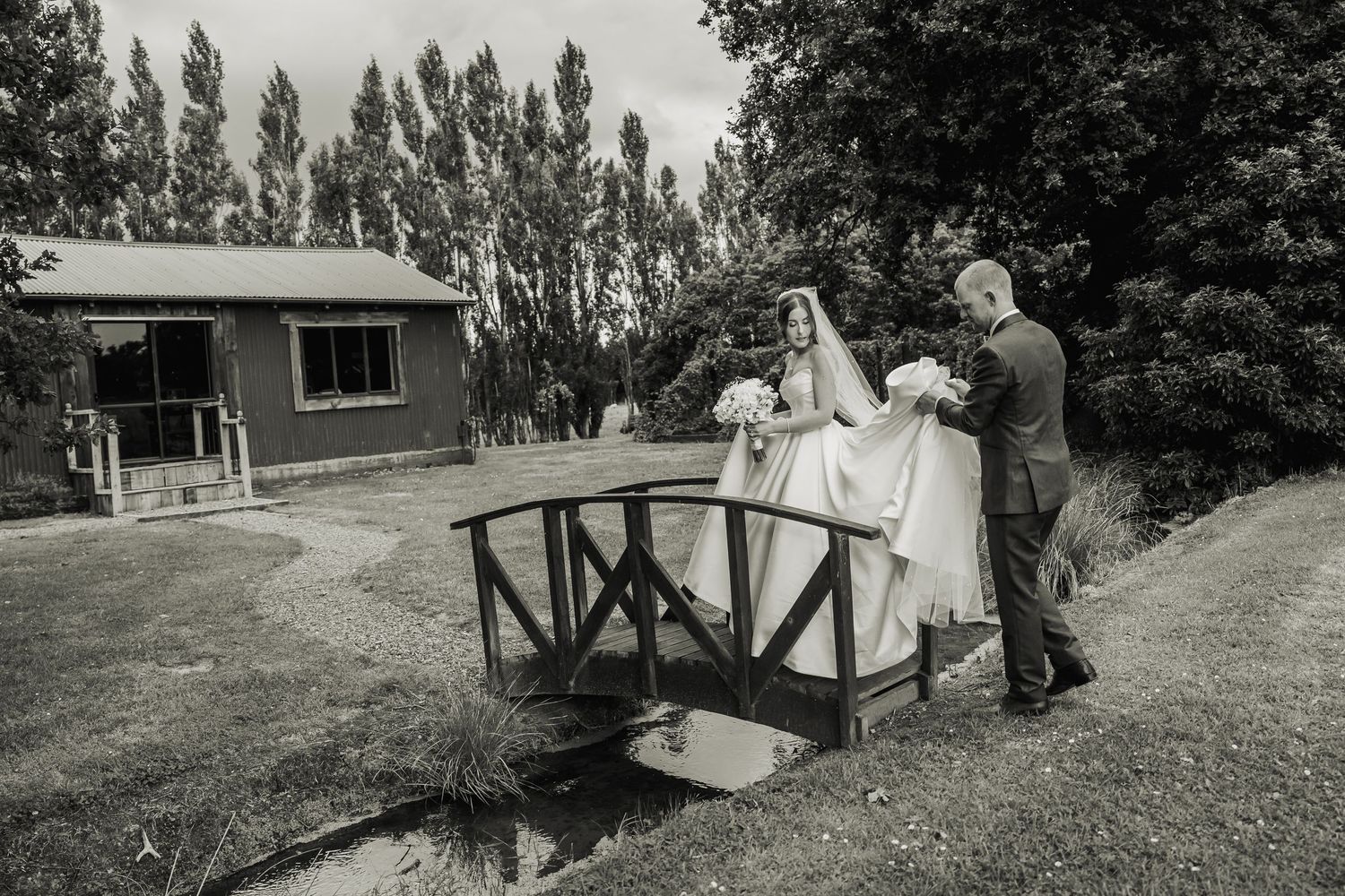 Groom helps bride cross a small wooden bridge in a black and white wedding photo.