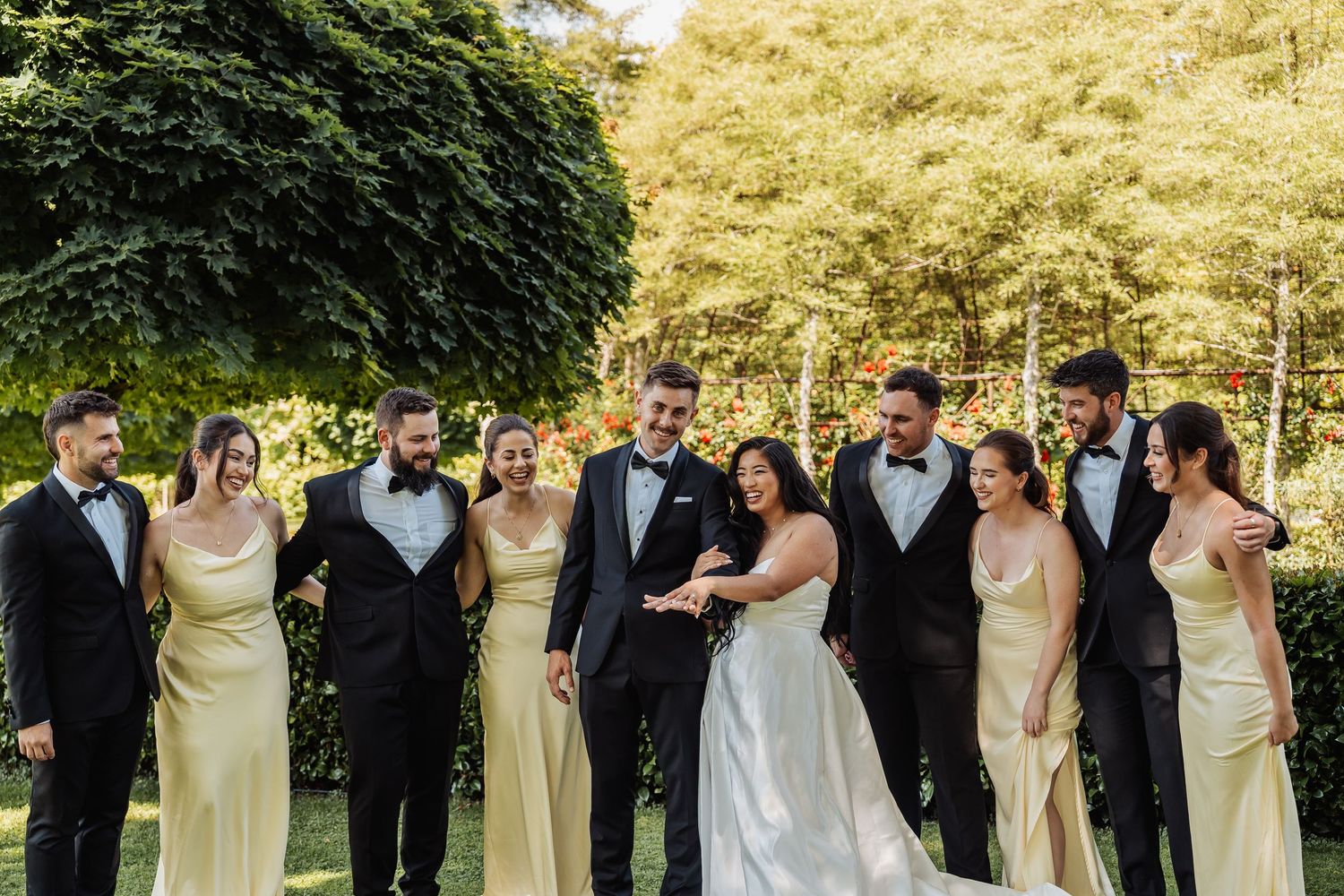 Wedding party in garden, bride and groom center, bridesmaids in yellow gowns and groomsmen in black tuxedos.