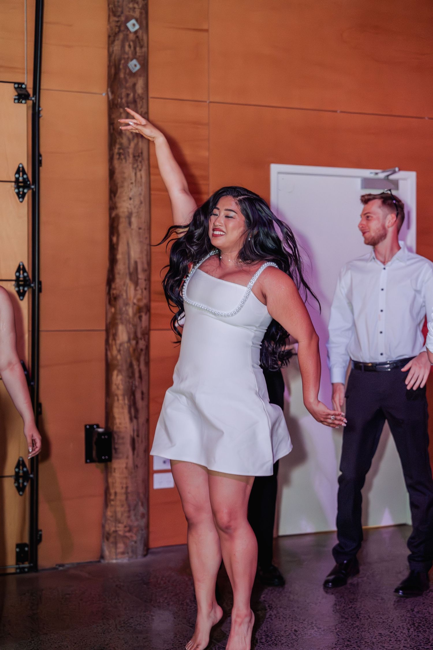 Bride in short white dress dances joyfully at wedding reception in warmly lit venue.