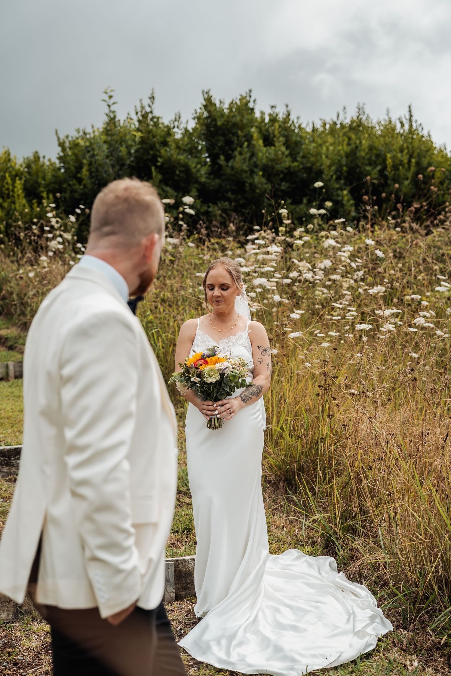 Bride in white gown holds colorful bouquet while groom in cream suit stands in foreground at outdoor wedding.