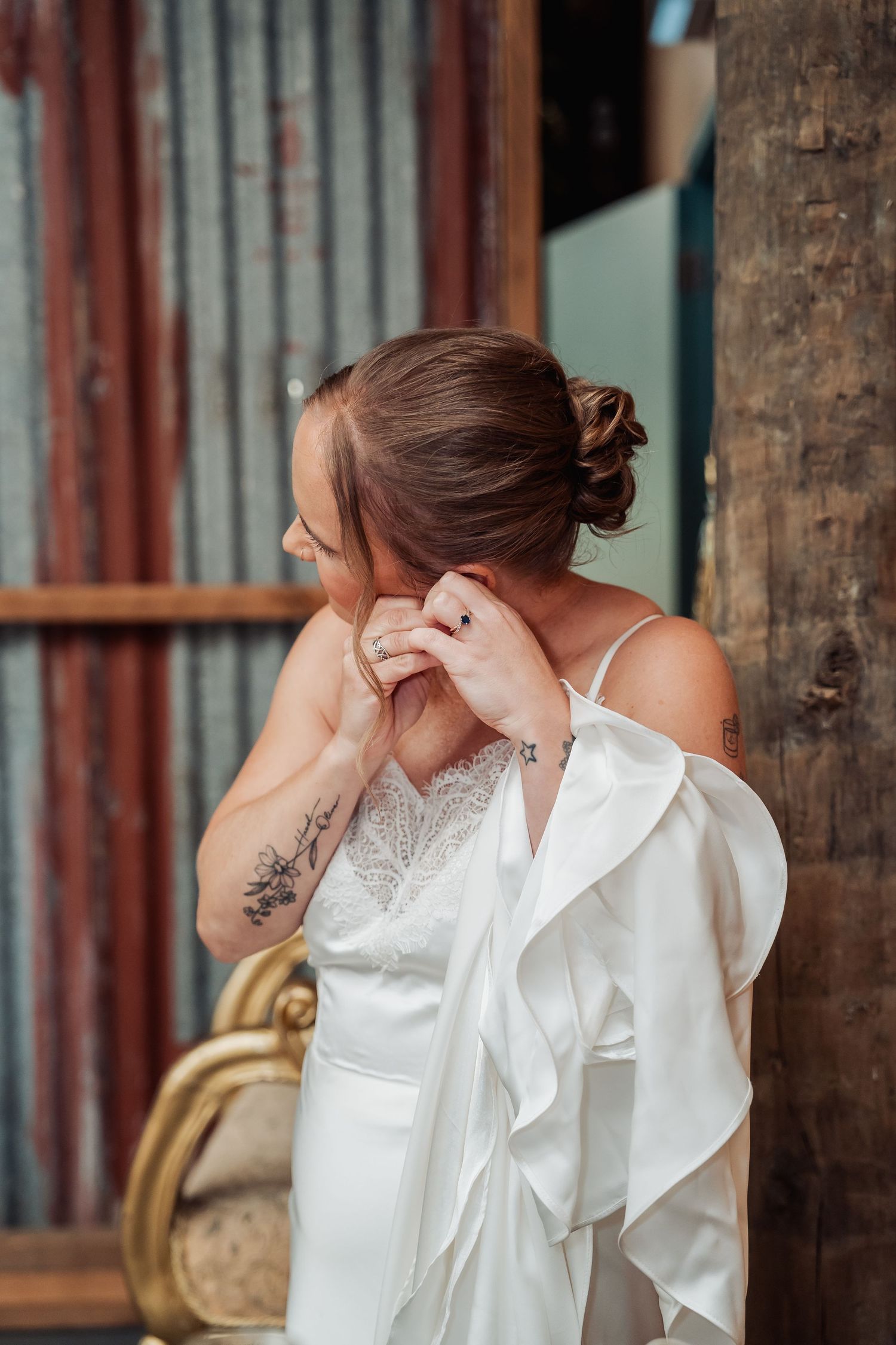 Bride in white satin gown adjusting earring, showing tattoos and elegant updo hairstyle.