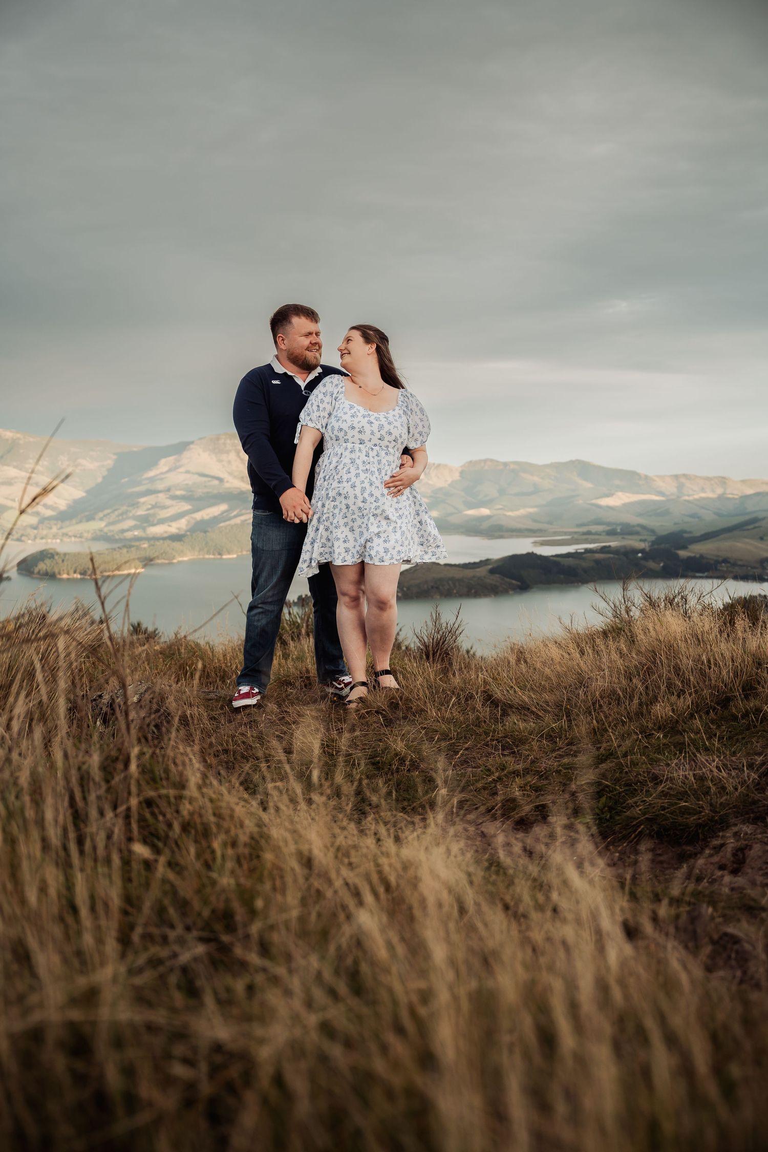Couple holding hands on a hilltop with stunning lake and mountain views in the background.