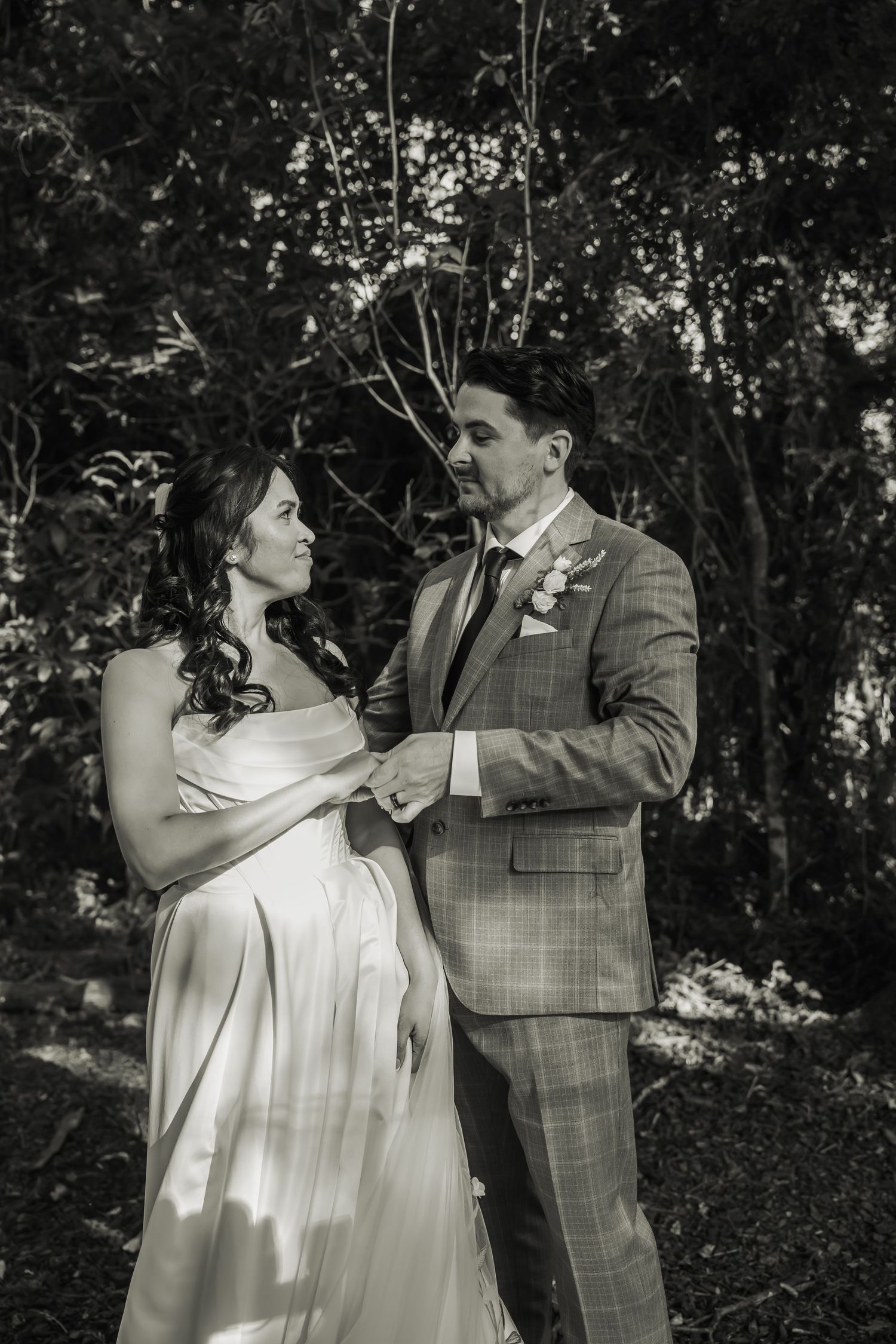 Black and white photo of a bride and groom holding hands, gazing at each other in a lush forest setting.