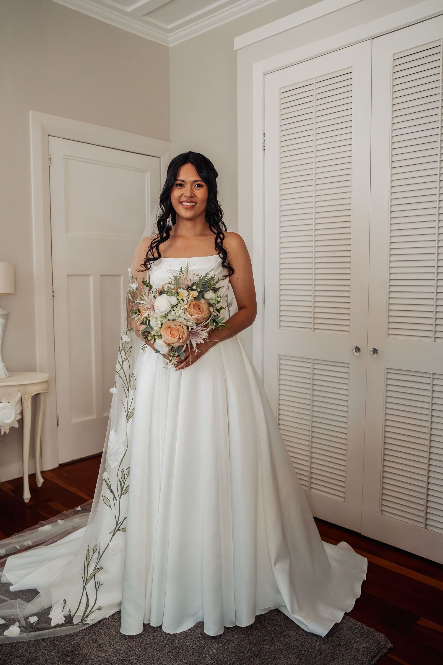 Bride in elegant white A-line wedding gown with floral veil holds peach bouquet indoors.