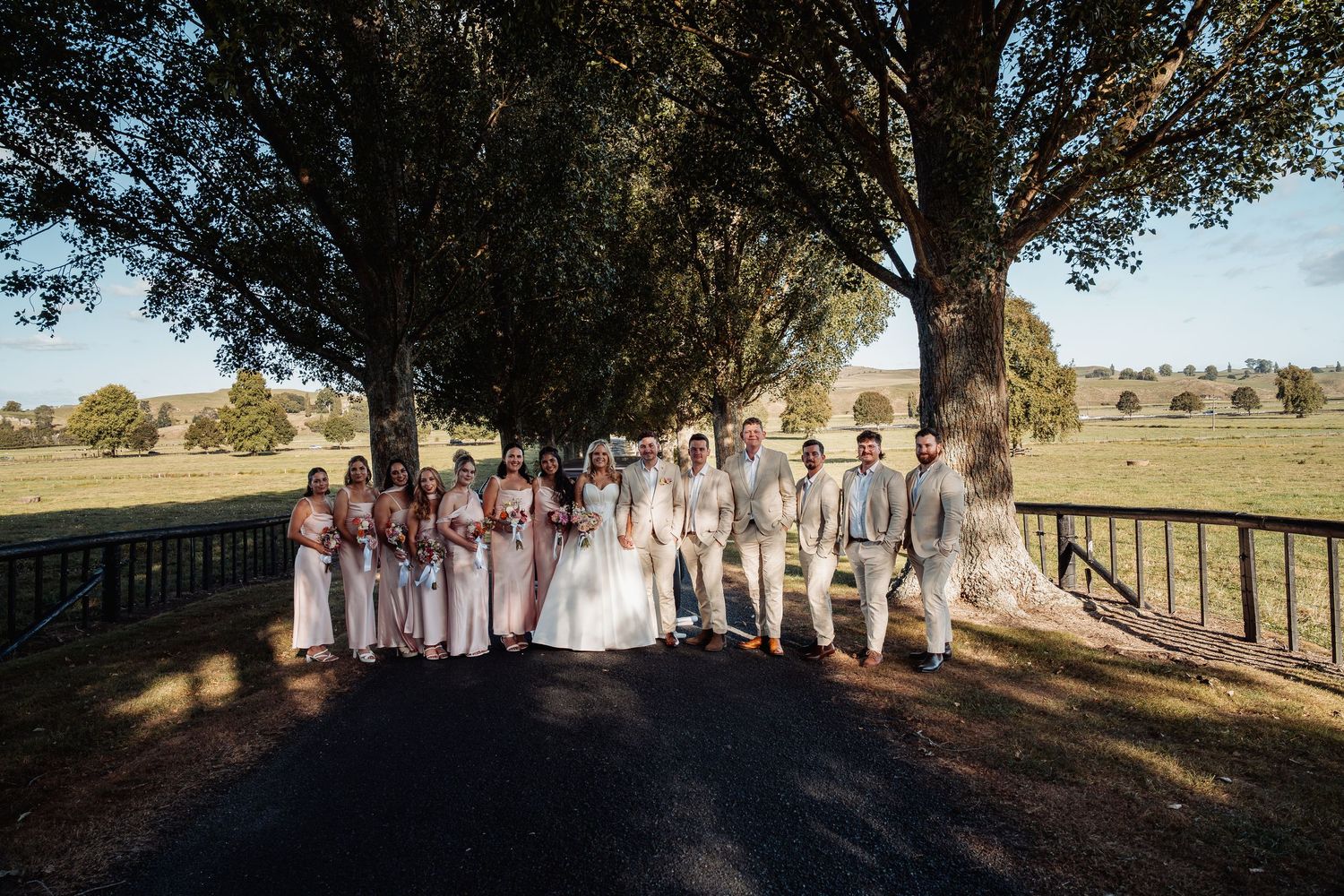 Wedding party in blush and beige attire poses along tree-lined driveway at rustic countryside venue.