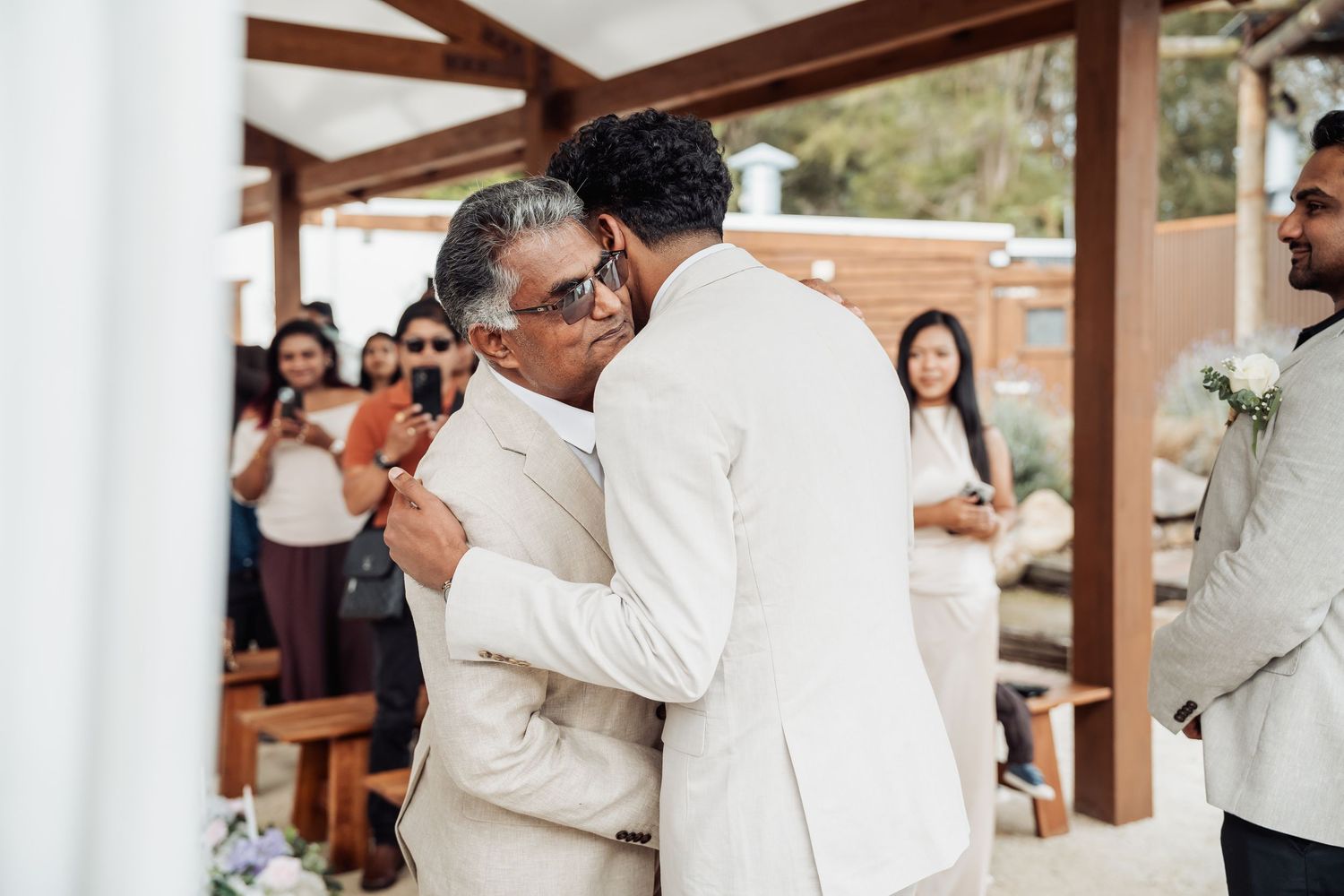 Groom in white suit sharing emotional hug with father at wedding ceremony.