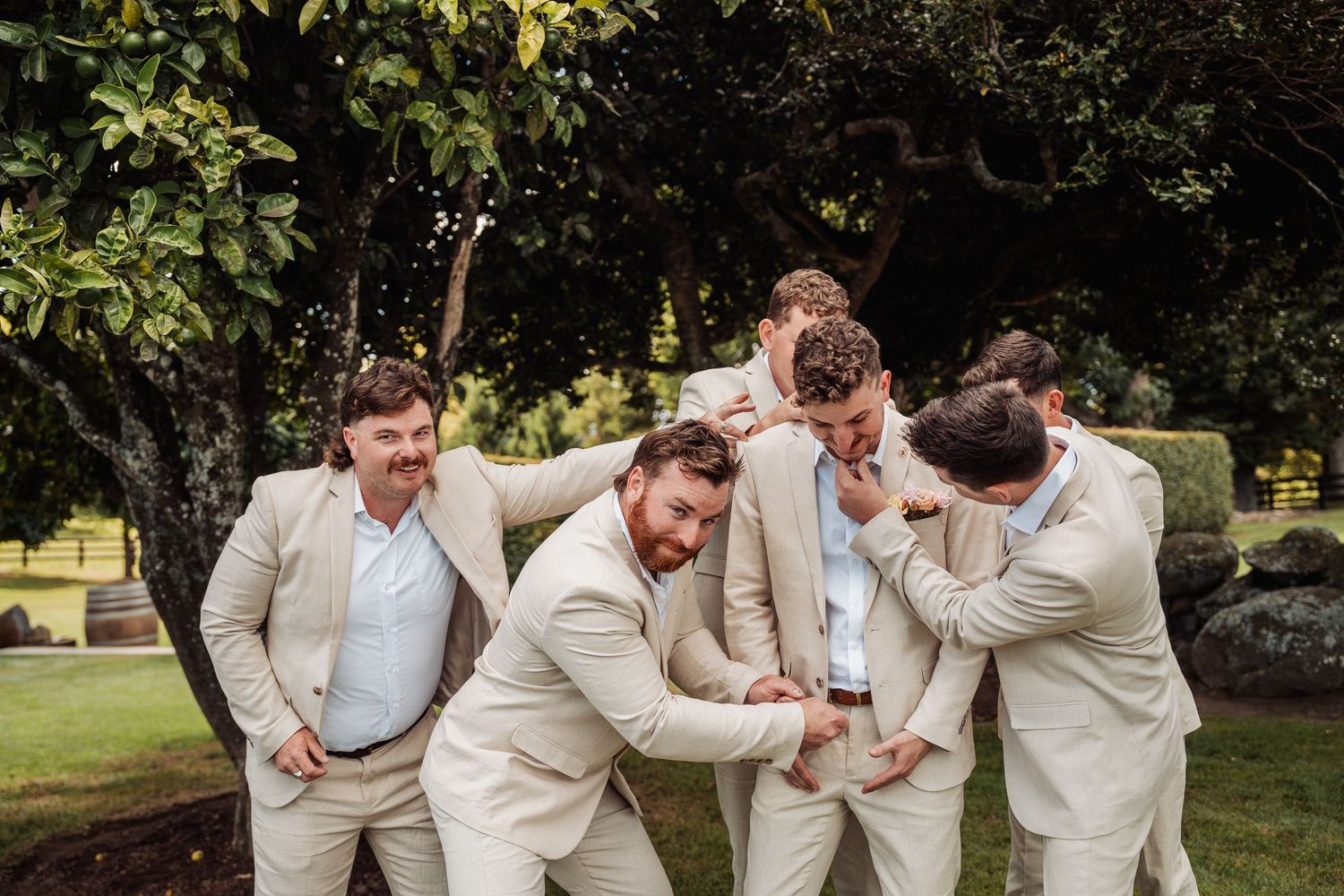 Groomsmen in tan suits laughing and joking together outdoors under large green trees.