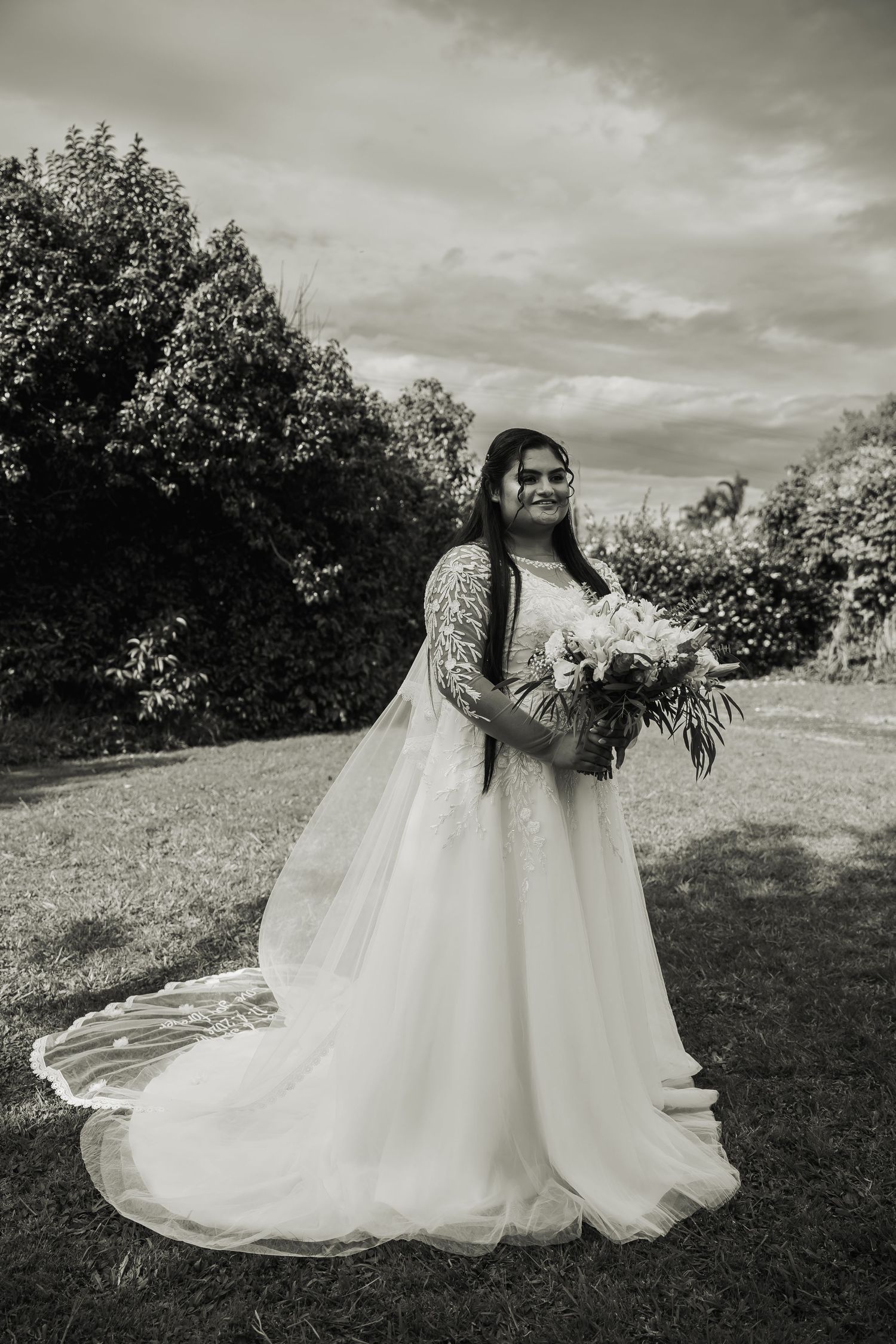Black and white portrait of smiling bride in lace ball gown holding bouquet in outdoor garden setting.