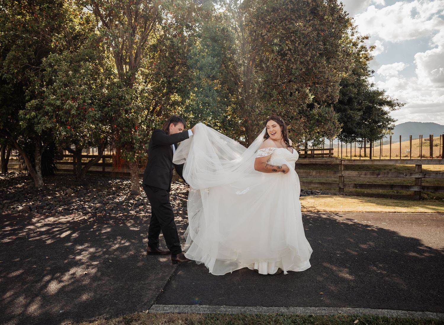 Groom in black suit lifts bride's veil as she smiles in white off-shoulder wedding gown outdoors.