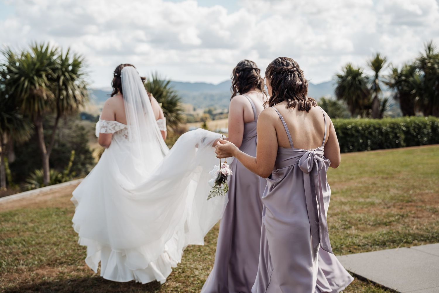 Bride in white gown and veil walks with two bridesmaids in lavender dresses on a green lawn with palm trees.