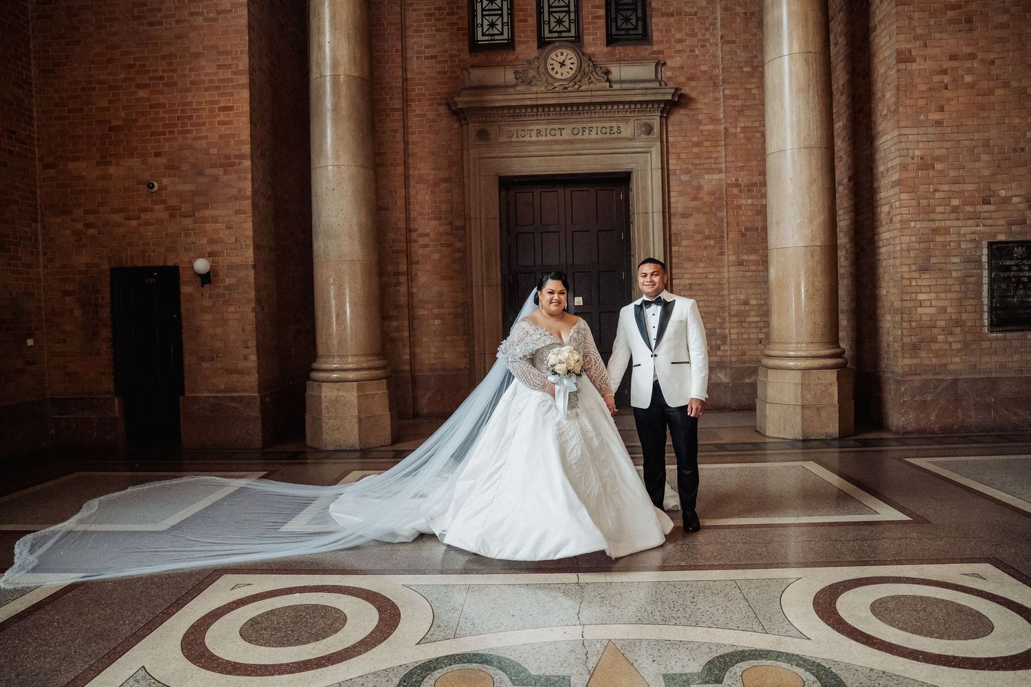 Wedding couple in white dress and tuxedo posing in front of ornate brick building with marble floor details.
