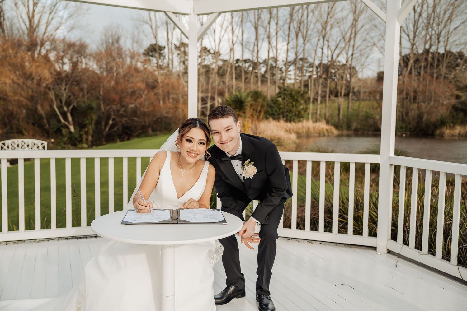 A couple sits together on a white gazebo bench surrounded by winter trees at their outdoor wedding venue.